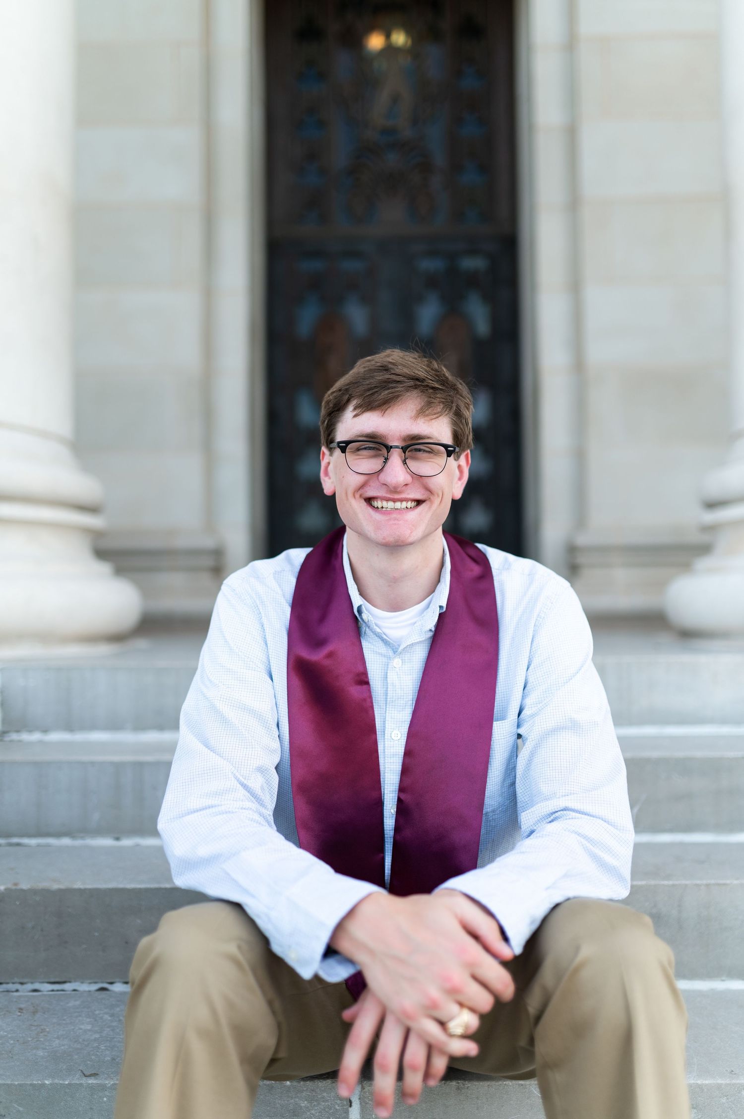 Smiling male Texas A&M grad in maroon stole sits on steps before ornate campus building doors. Grad Photos at the Texas A&M Admin building