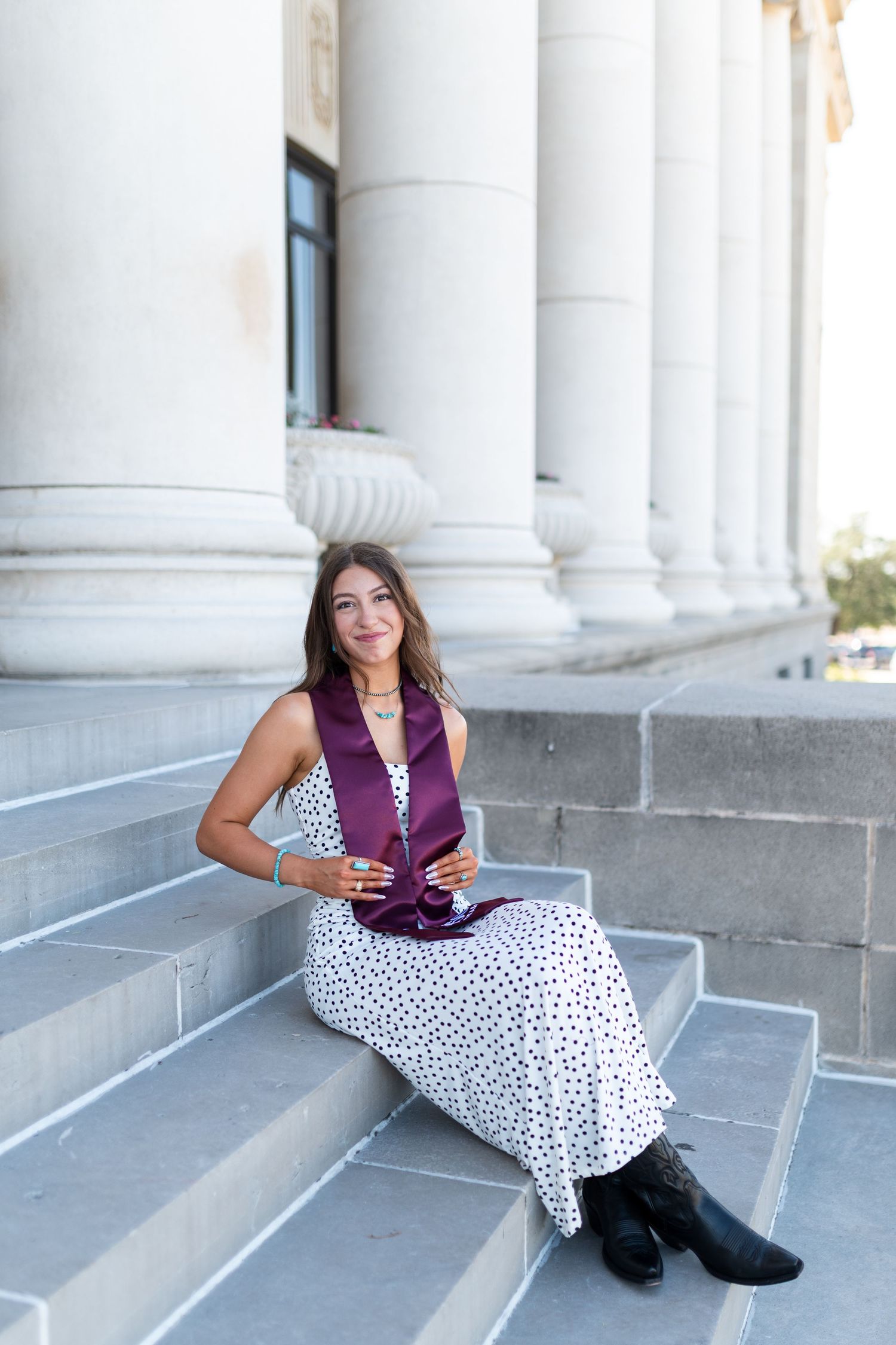 Smiling graduate in white polka dot dress and maroon stole sits on stone steps by columns. Grad Photos at the Texas A&M Admin building