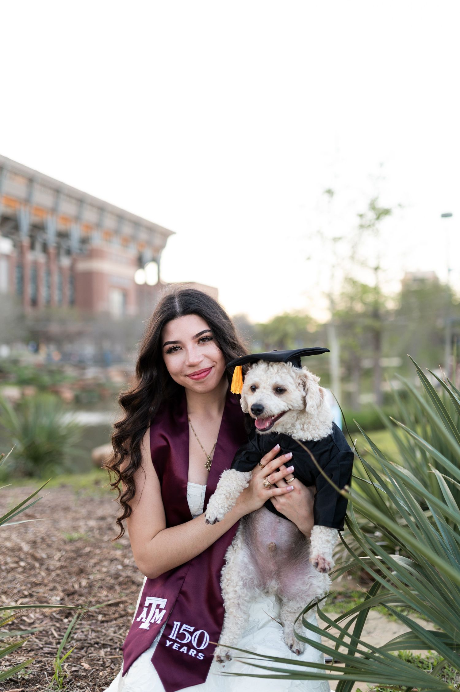 Young woman in white dress and Texas A&M sash holds dog in graduation attire on campus grounds.