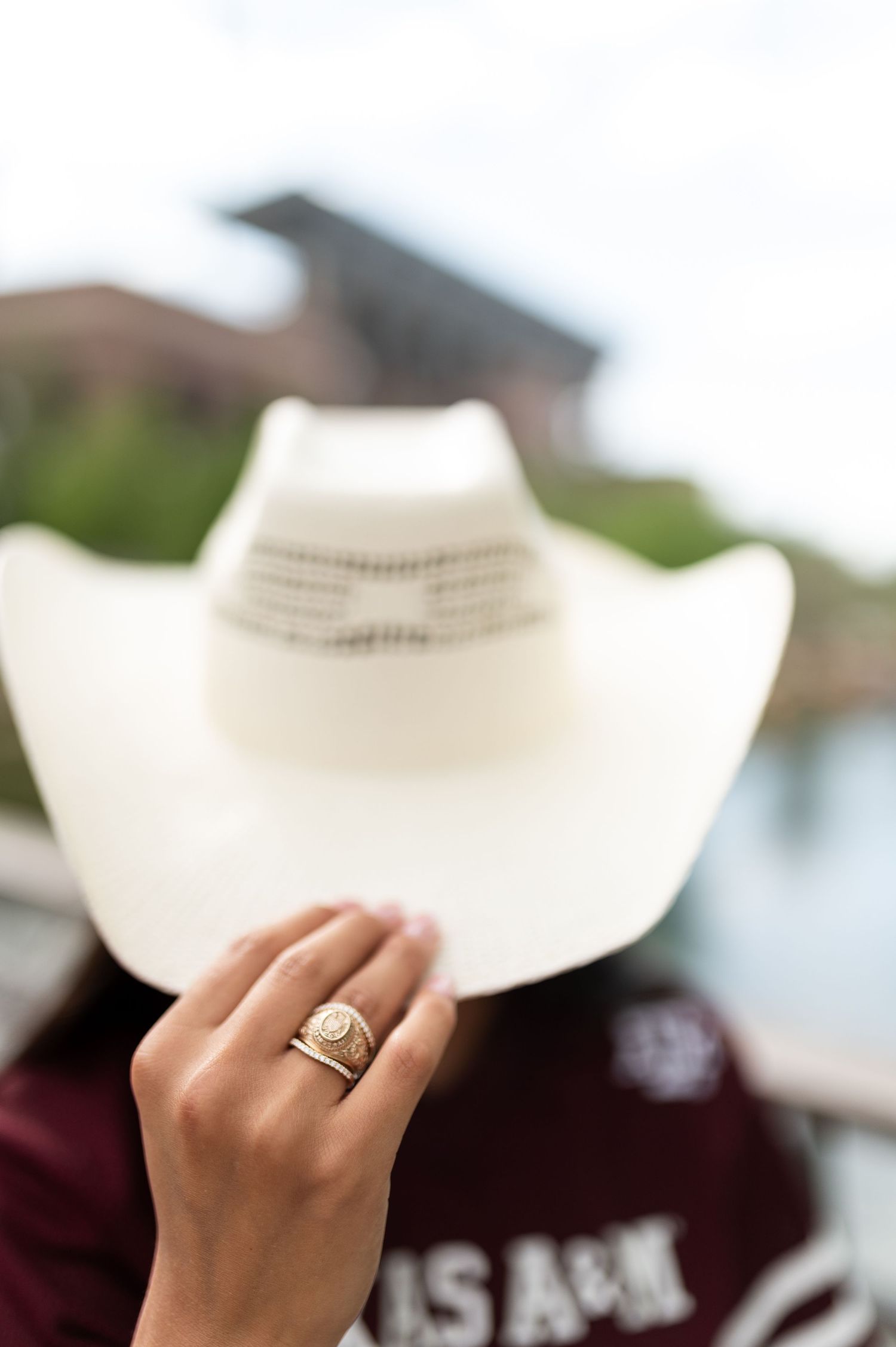 A hand displaying a gold ring held up in front of a white cowboy hat outdoors.