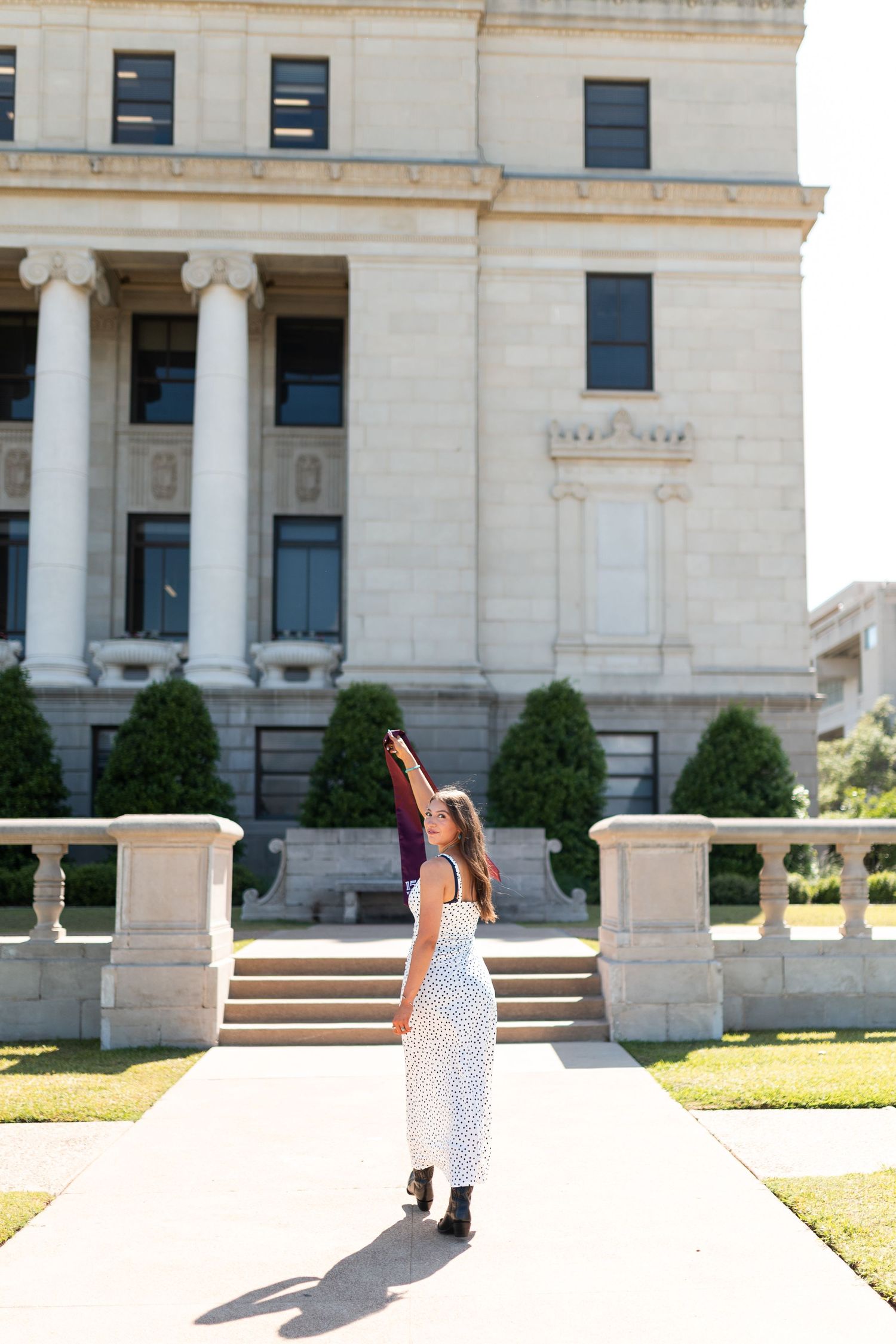 Female Texas A&M graduate raises maroon stole triumphantly outside neoclassical university building on sunny day. Grad Photos at the Admin