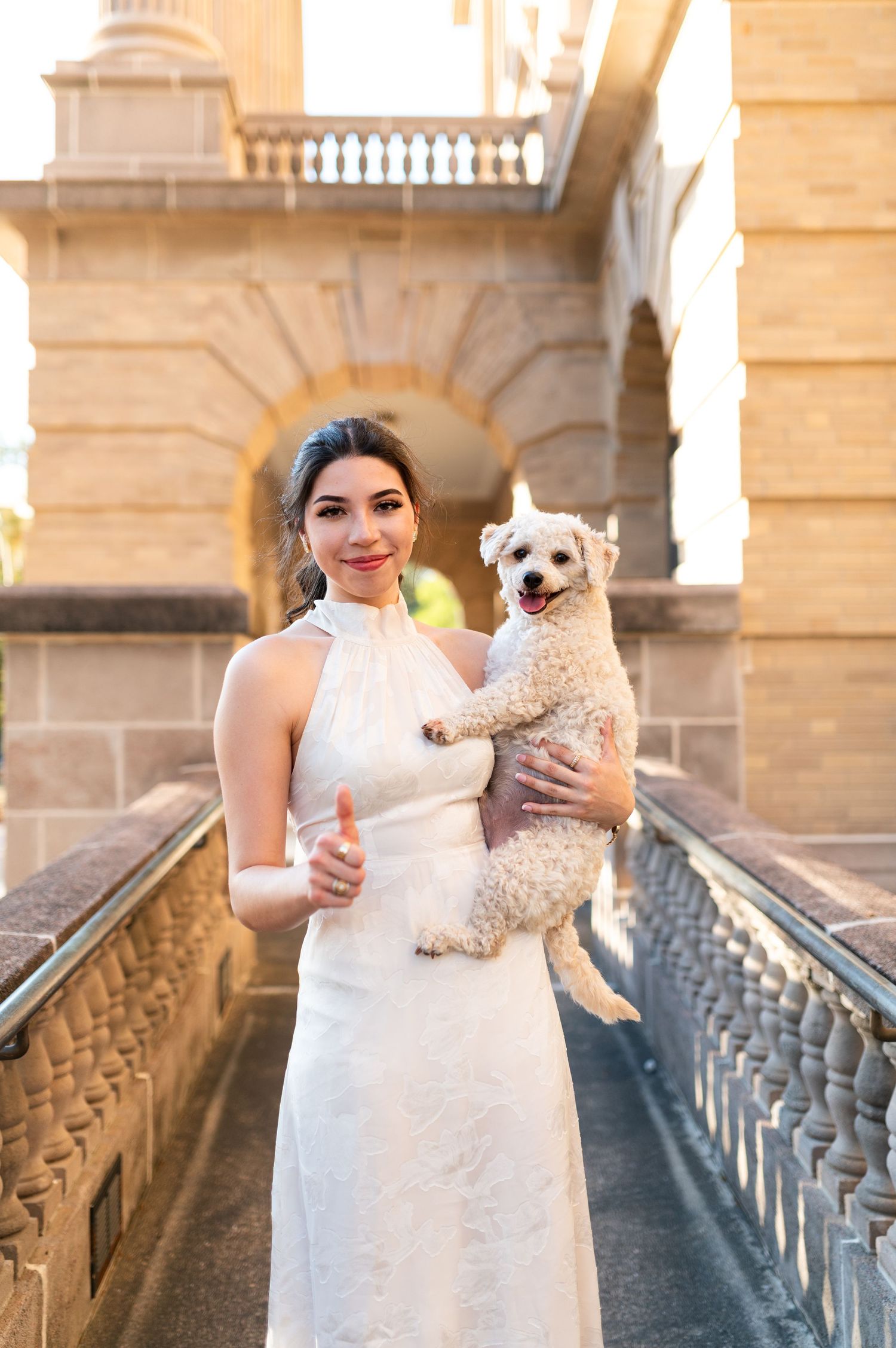 Smiling woman in white halter dress holds fluffy white dog outside historic building.