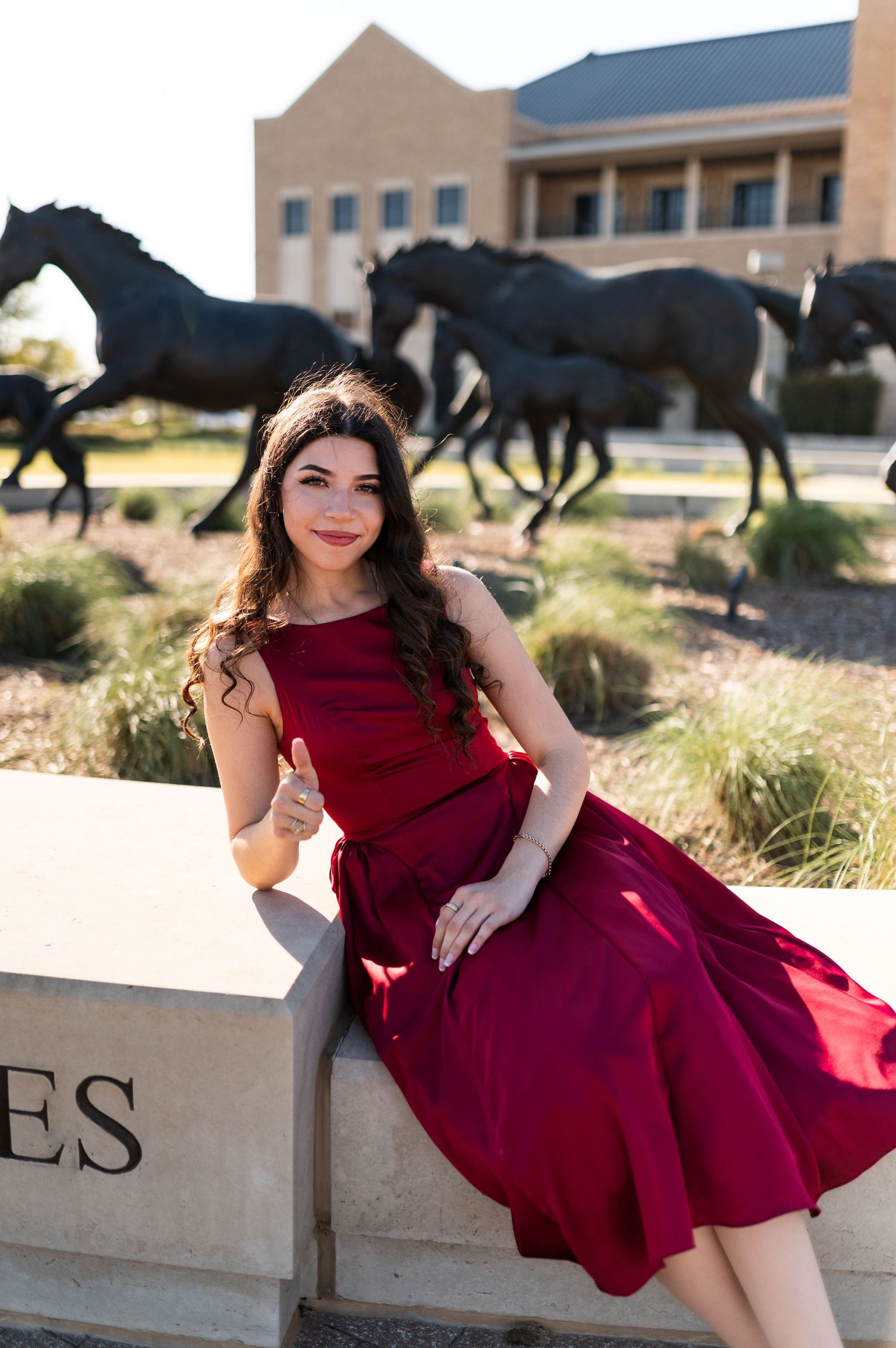 Woman in maroon dress poses on ledge before bronze horse sculptures at Texas A&M campus.