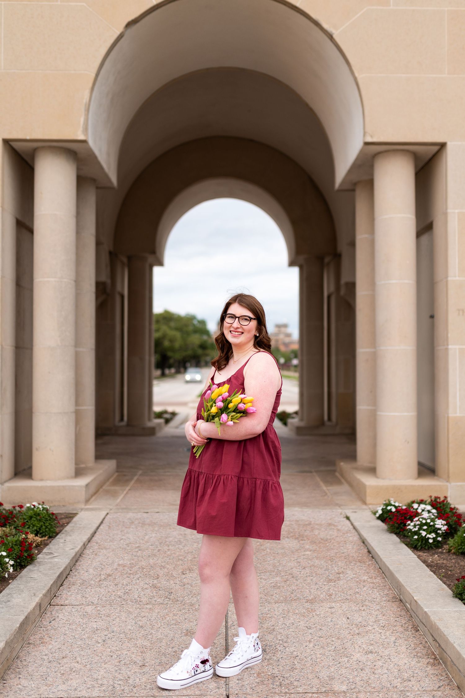 Texas A&M graduate in maroon dress holding colorful tulips under stone archway on campus.