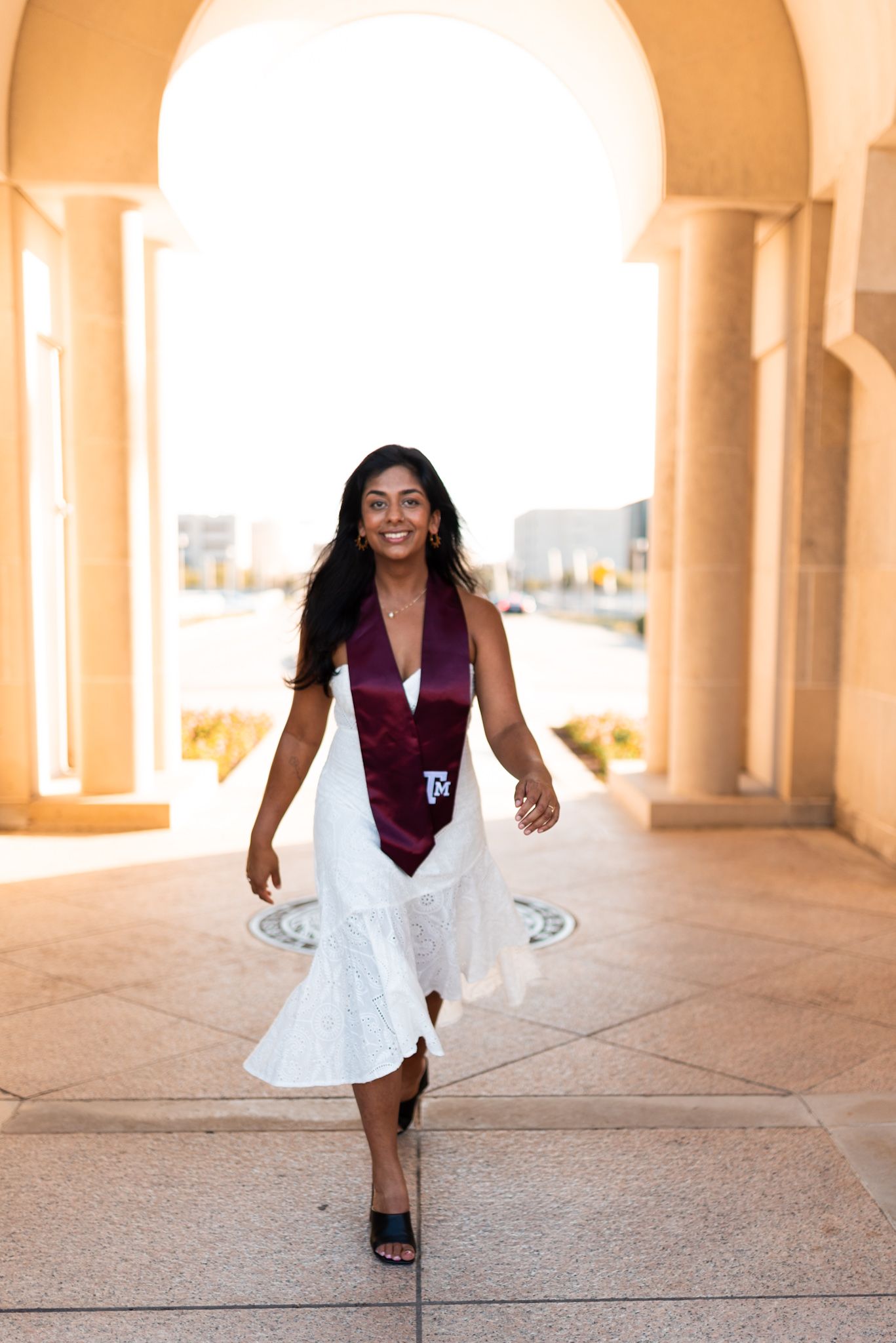 Smiling graduate in white dress and maroon Texas A&M stole walking under archway.