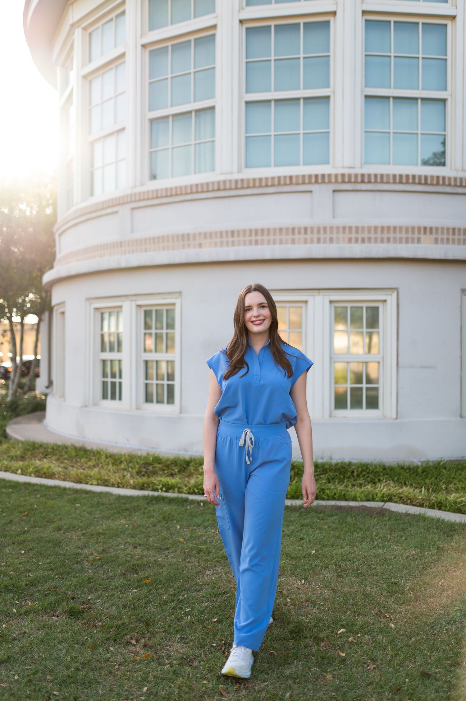 Smiling young woman in blue jumpsuit walks on green lawn in golden hour sunlight near white building.
