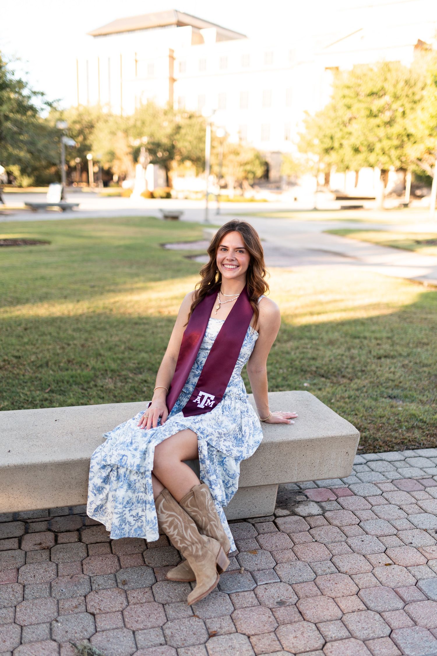 Texas A&M graduate in blue floral dress and cowboy boots sits on stone bench wearing maroon stole.