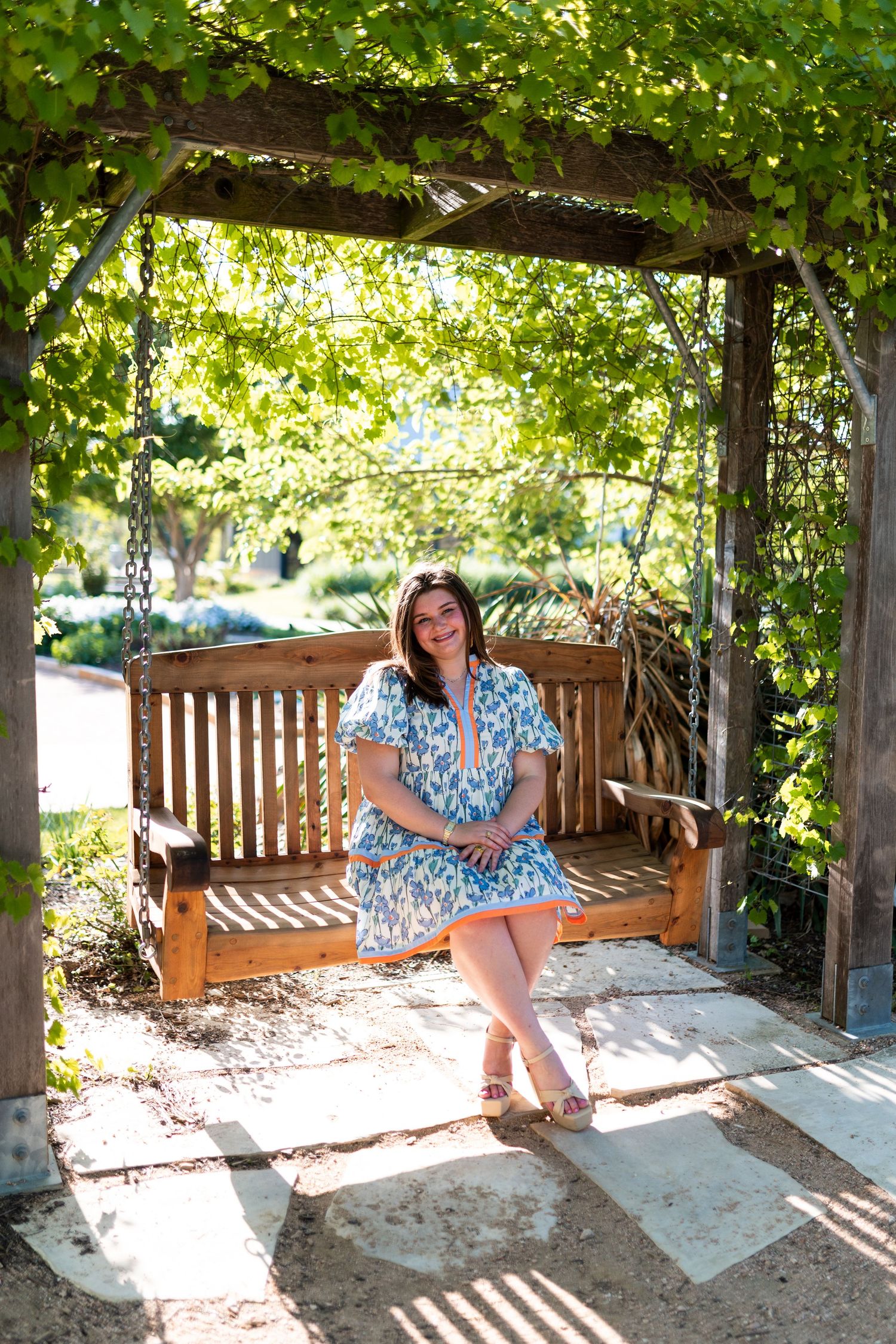 Woman in blue floral dress sitting on wooden garden swing under green vine-covered pergola.