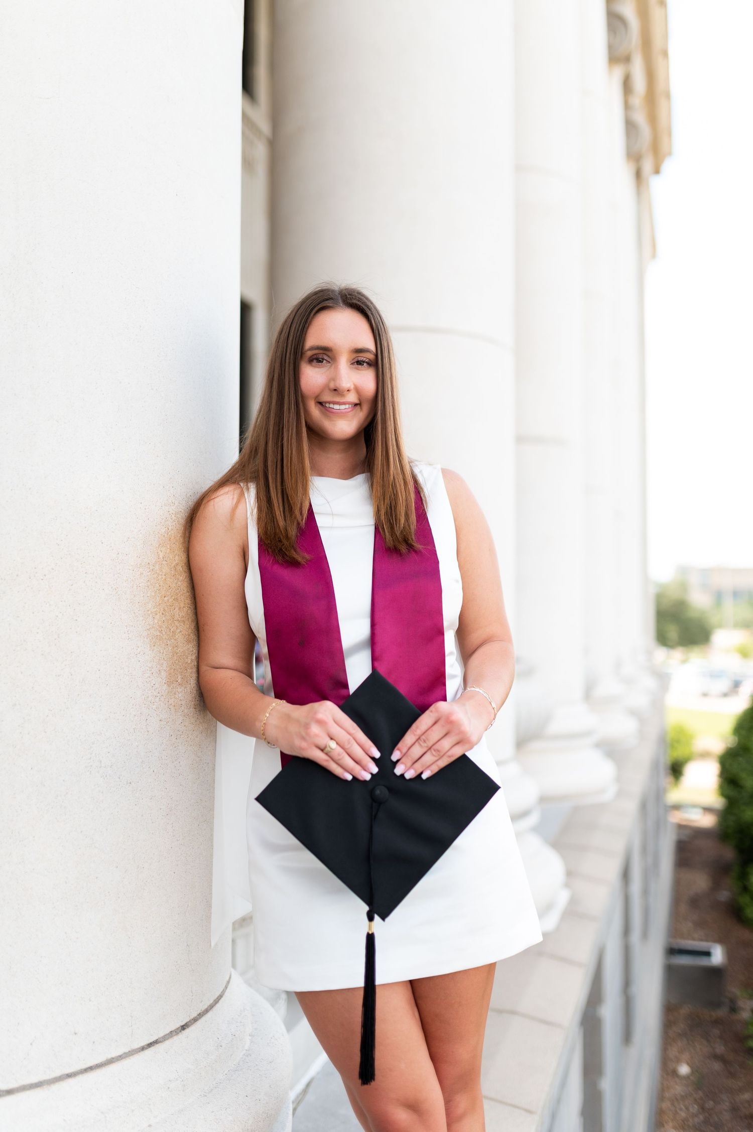 Smiling female graduate leans against column holding black cap with maroon stole. Grad Photos at the Texas A&M Admin building