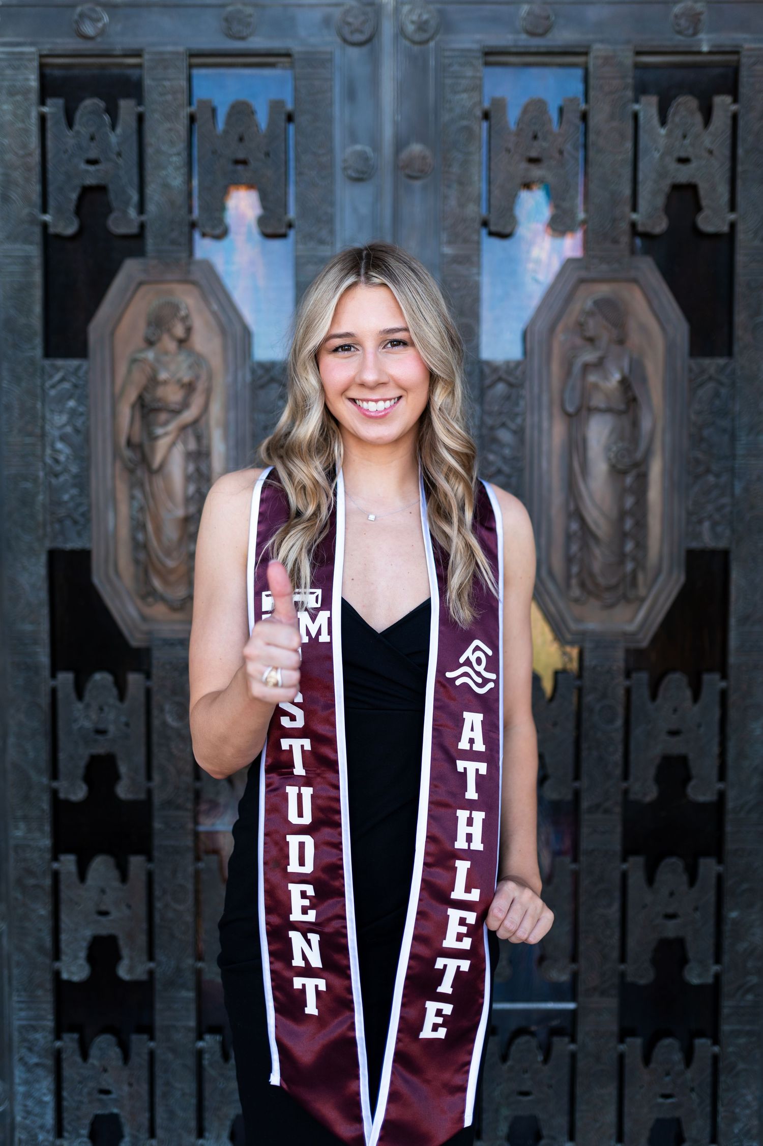 Smiling blonde woman displaying Texas A&M Student Athlete graduation stole before ornate doors. Grad Photos at the Admin building