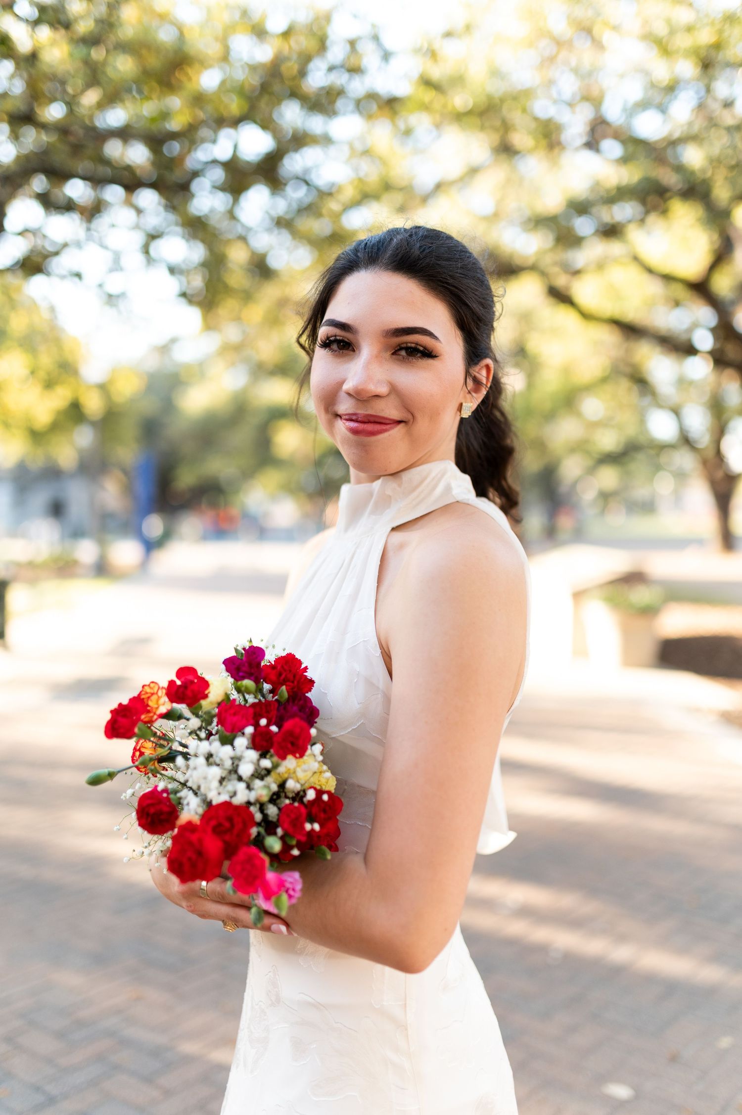 Smiling bride in white halter dress holds vibrant red and pink bouquet outdoors under oak trees.