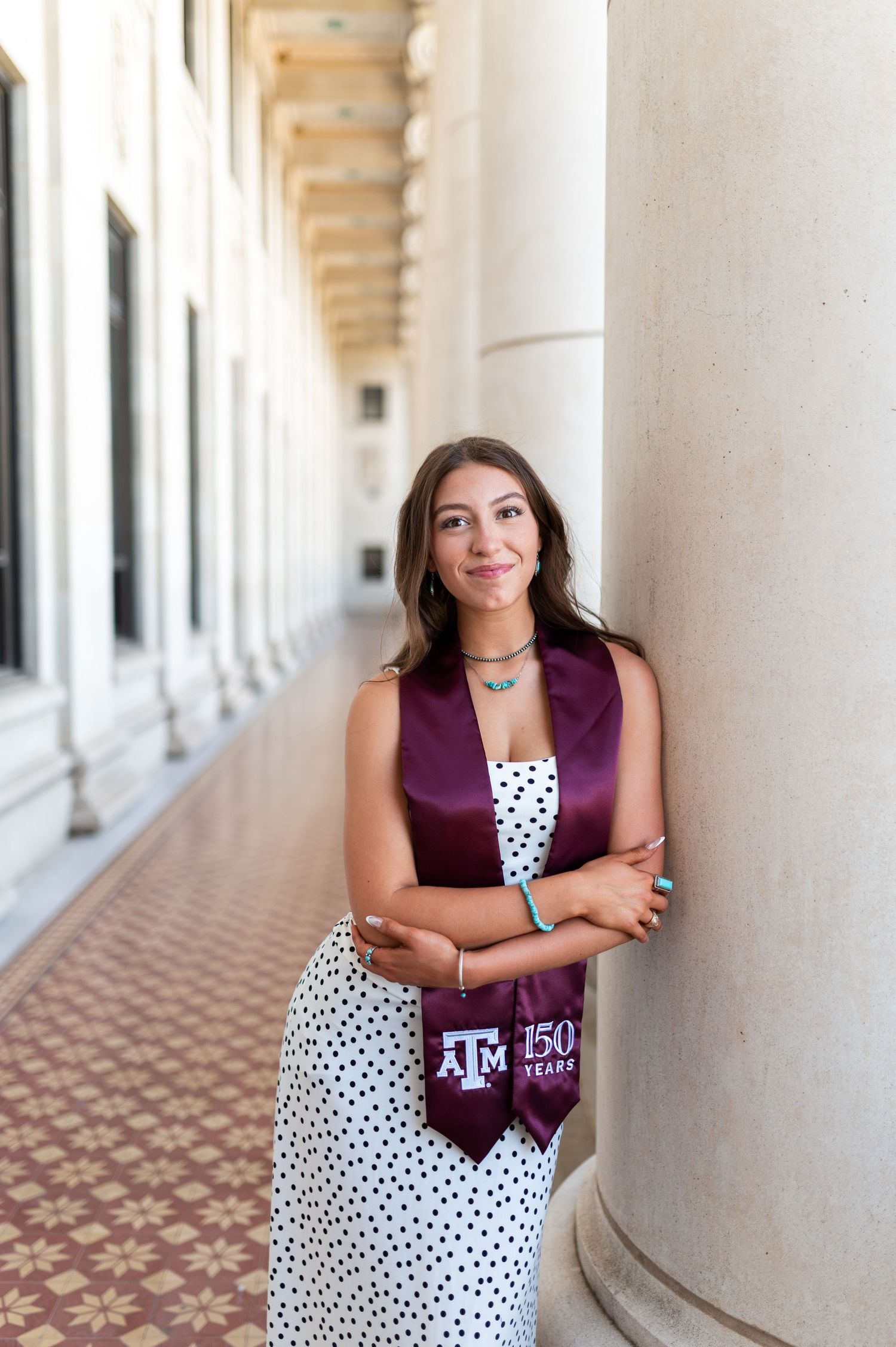 Smiling Texas A&M graduate in maroon stole leaning against column in elegant columned hallway. Grad Photos at the Texas A&M Admin building