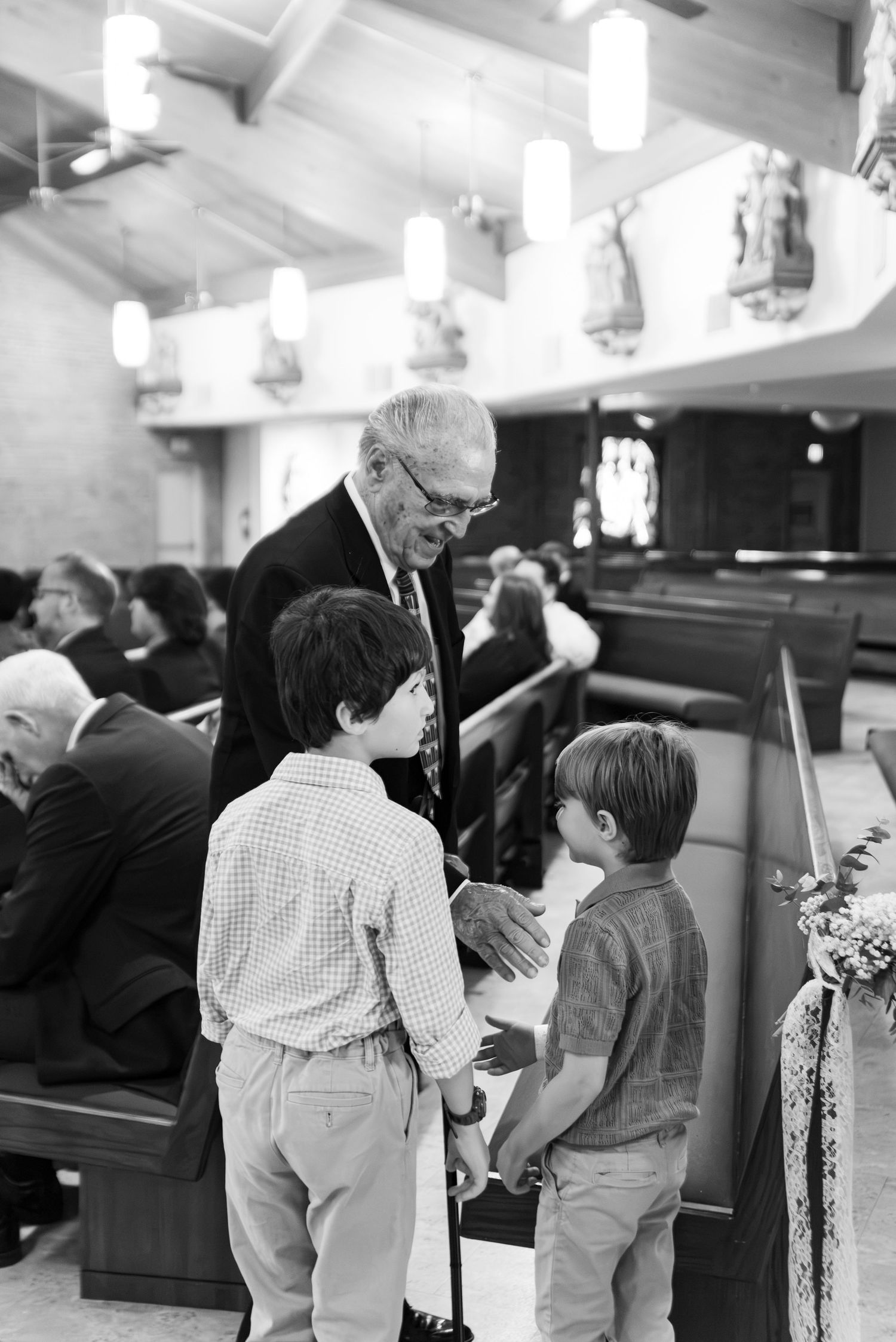 Black and white photo of elderly man greeting two young boys in church pew aisle.