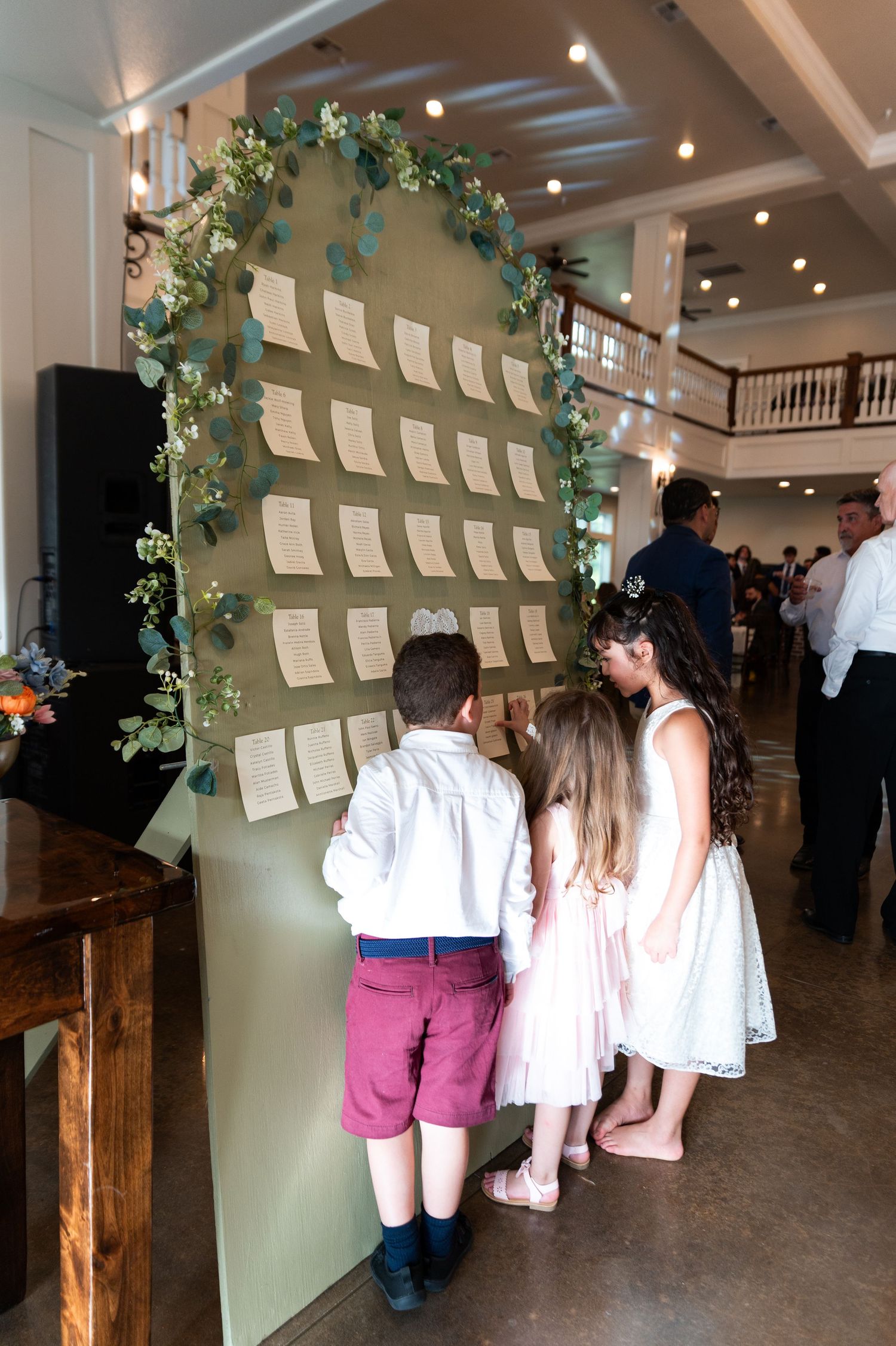 Children in wedding attire browse elegant greenery-adorned seating chart display at reception venue entrance.