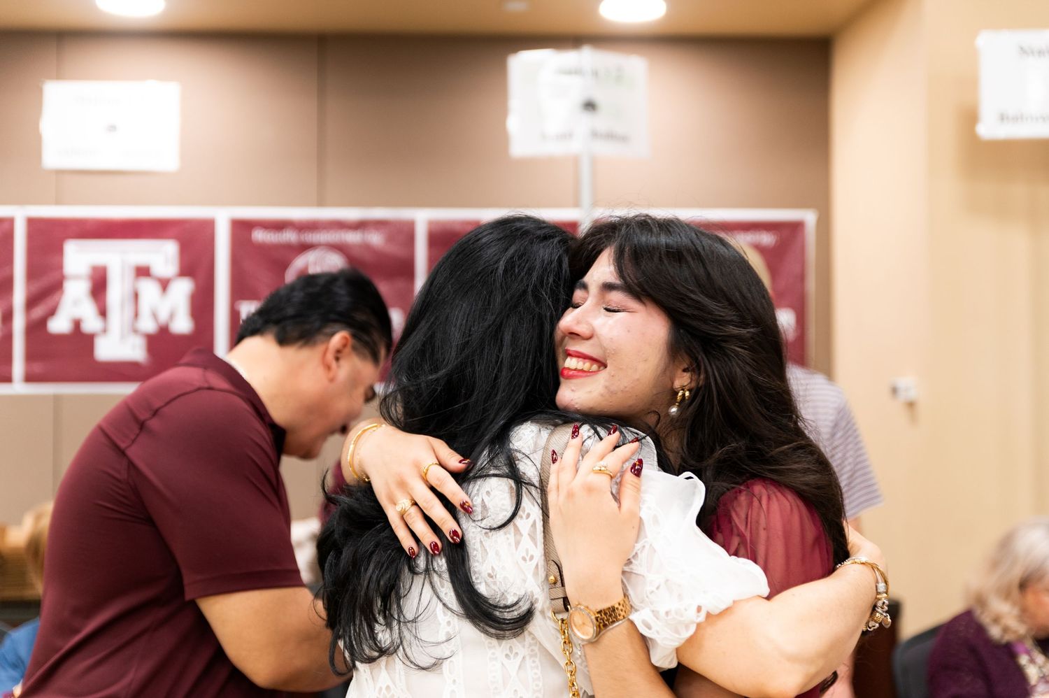 Two women share a warm hug at a Texas A&M University event, smiling joyfully in a maroon-decorated room.