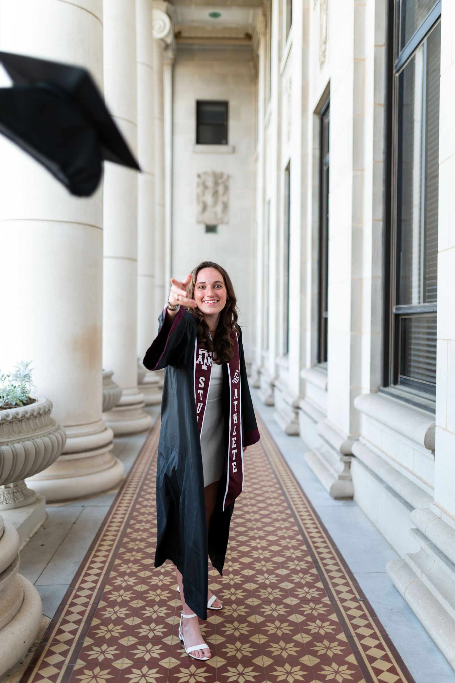 Joyful Texas A&M graduate tosses cap while walking in graduation gown through campus corridor. Grad Photos at the Admin building