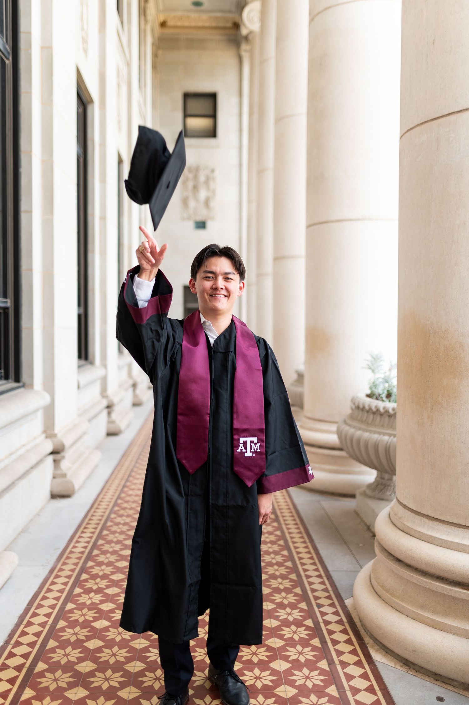 Texas A&M graduate in black gown tosses cap while making peace sign in campus corridor. Grad Photos at the Admin building