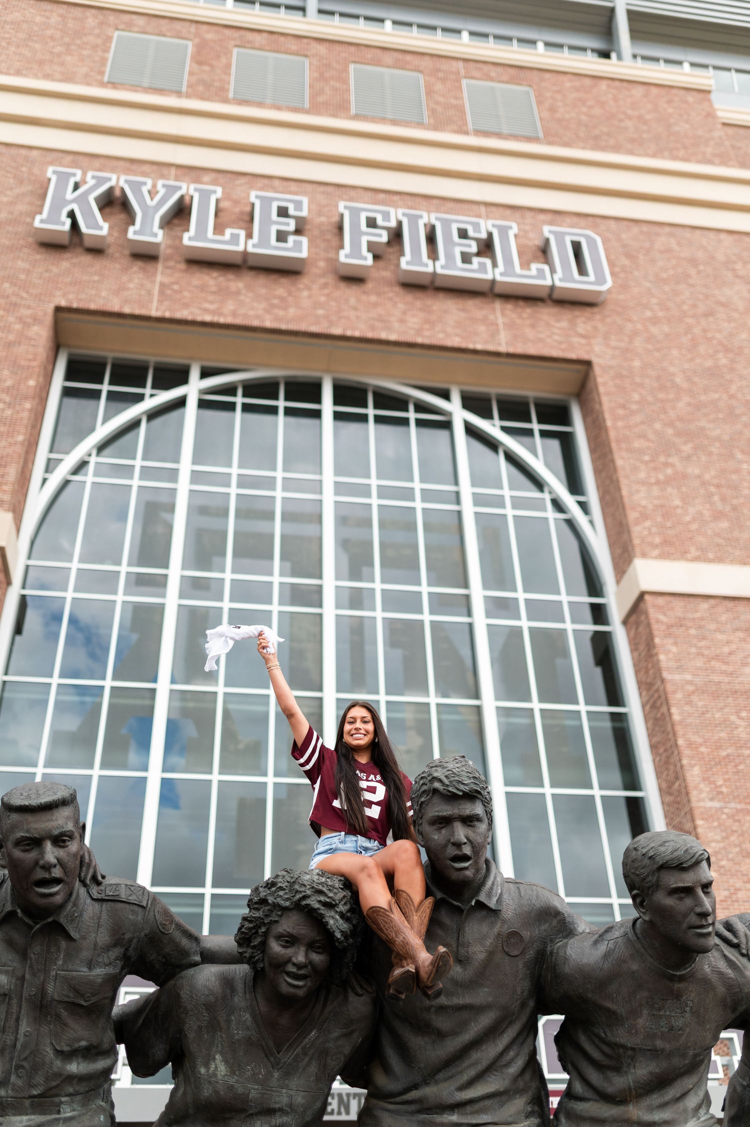 Smiling woman in Texas A&M jersey perches on bronze fan statues outside Kyle Field.