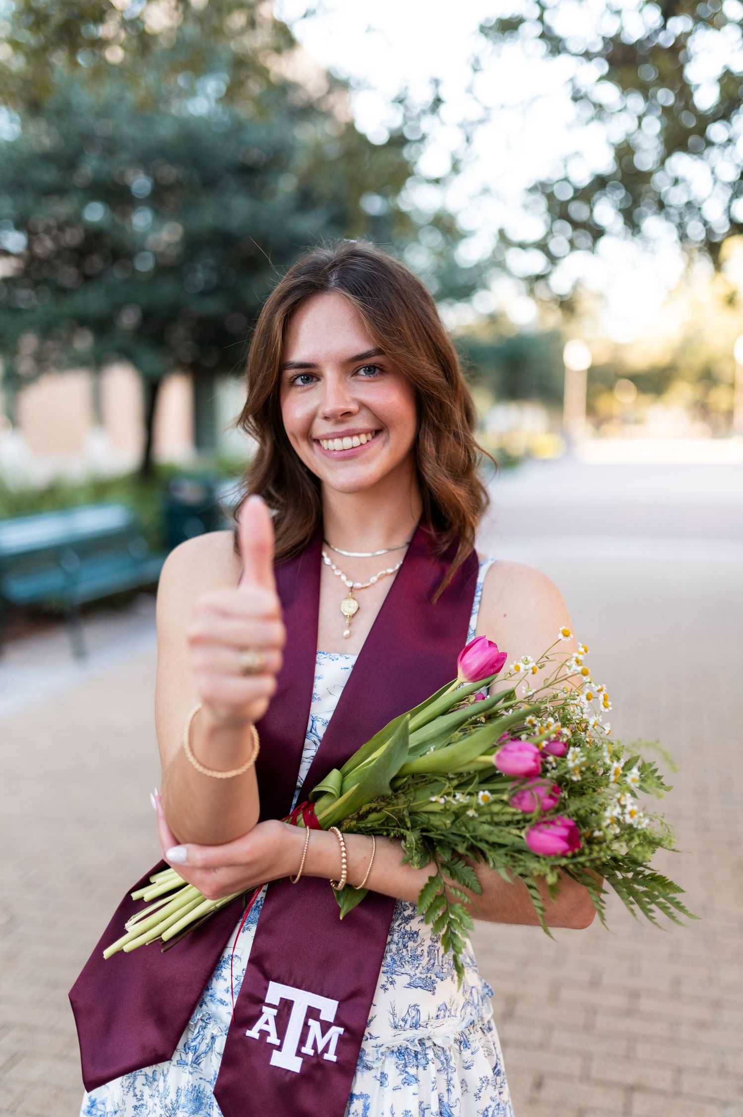 Smiling Texas A&M graduate in maroon stole gives thumbs up while holding pink tulip bouquet outdoors.
