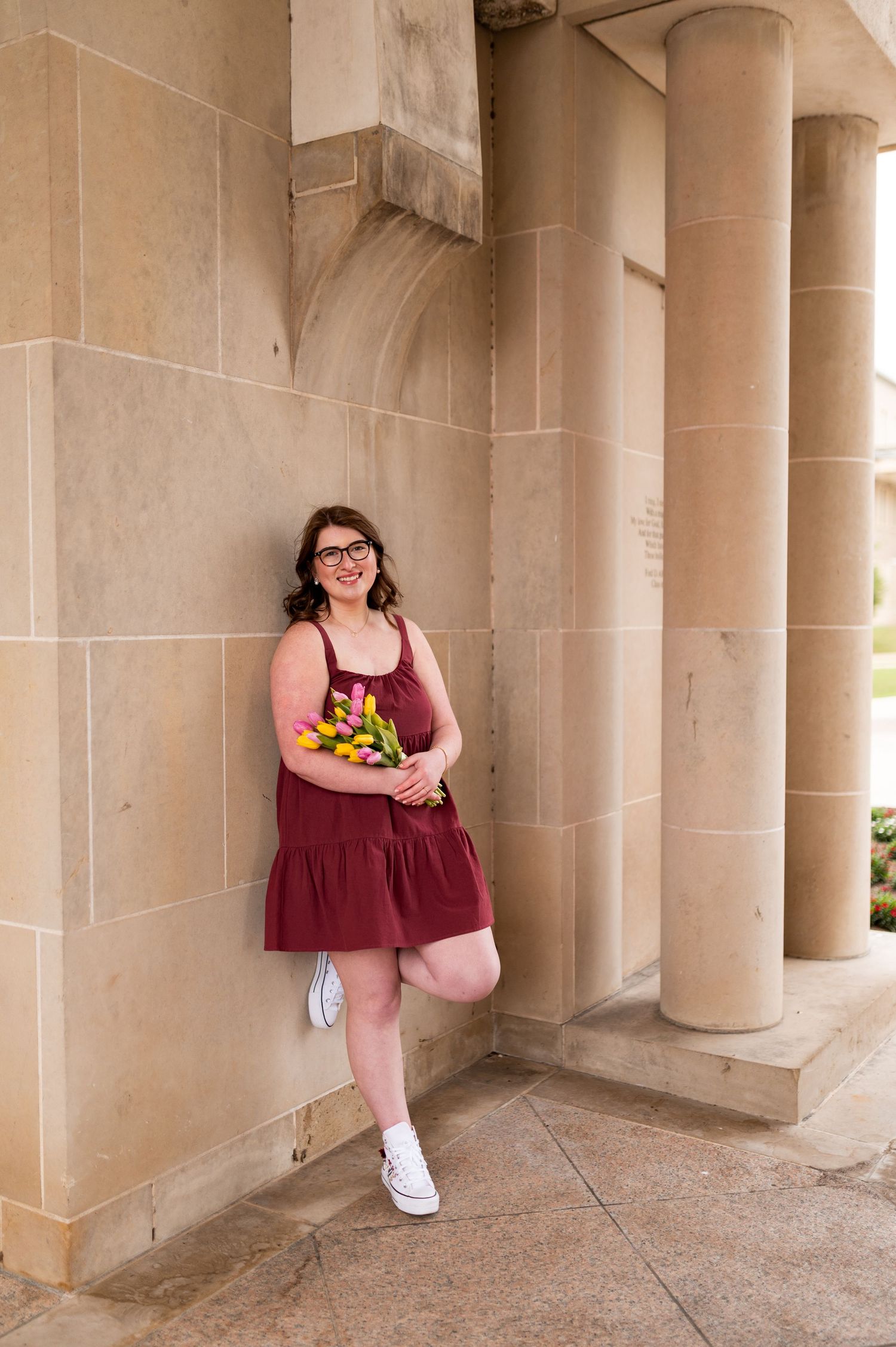 Young woman in maroon dress holding colorful tulips leaning against stone column building.