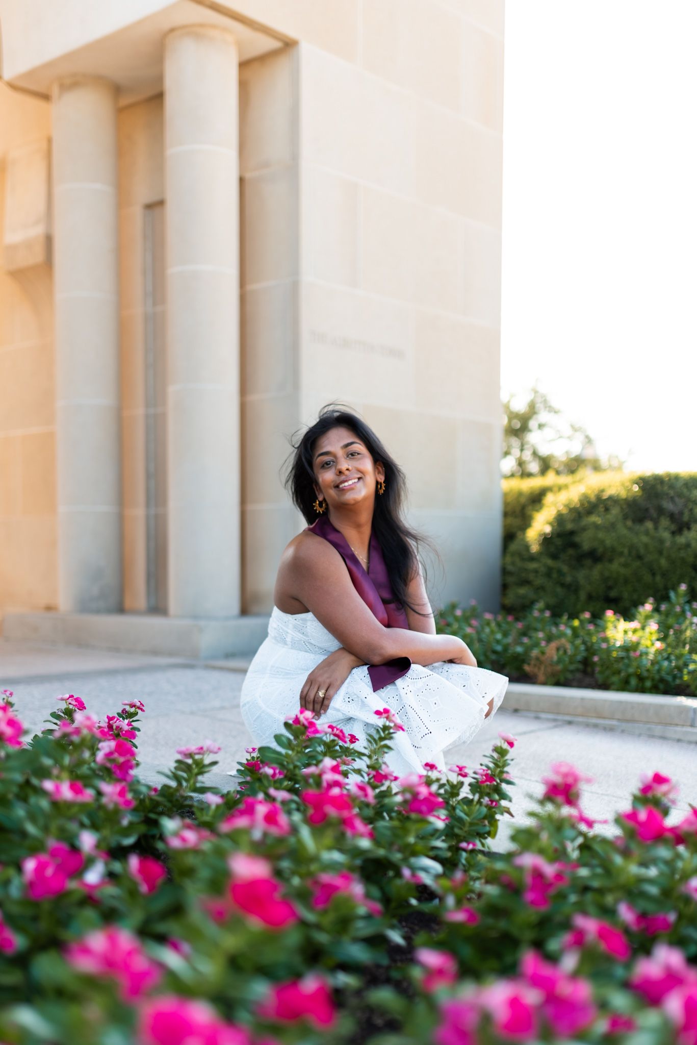 Smiling young woman in white dress sitting near vibrant pink flowers by stone columns.