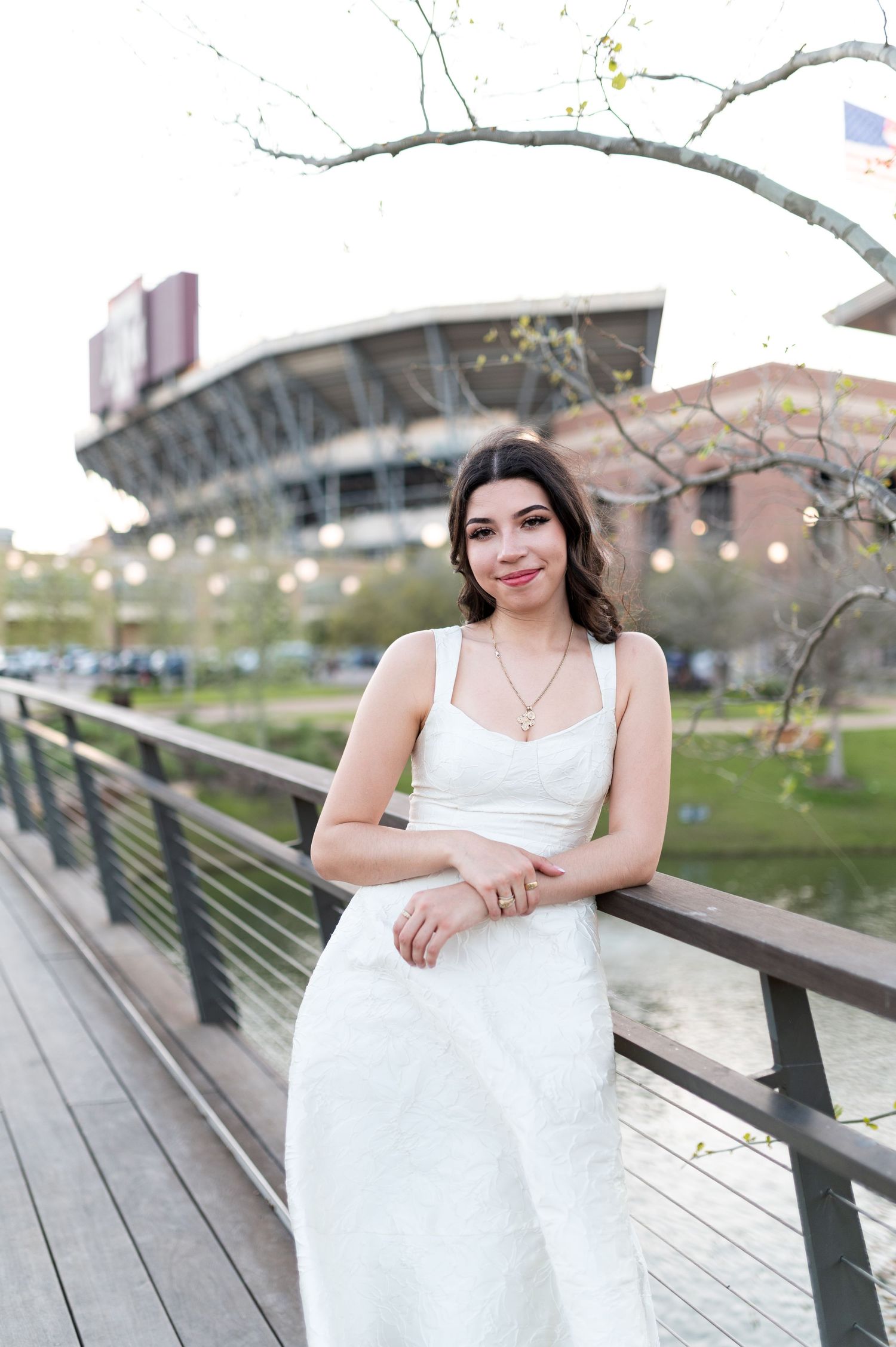 Young woman in white dress leans on wooden bridge railing with Texas A&M stadium in background.
