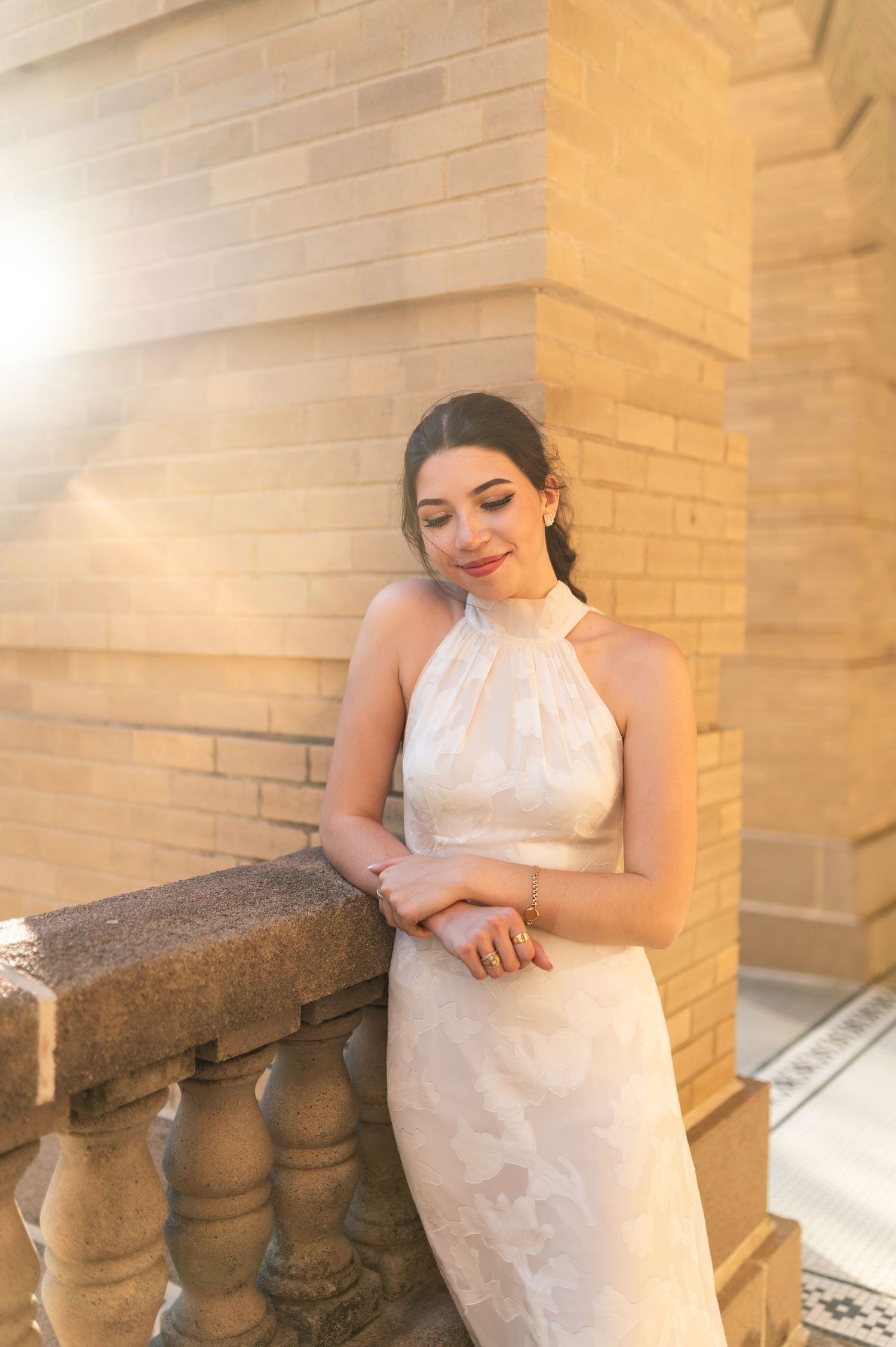 Woman in white halter gown leans on stone balustrade in golden sunlight.