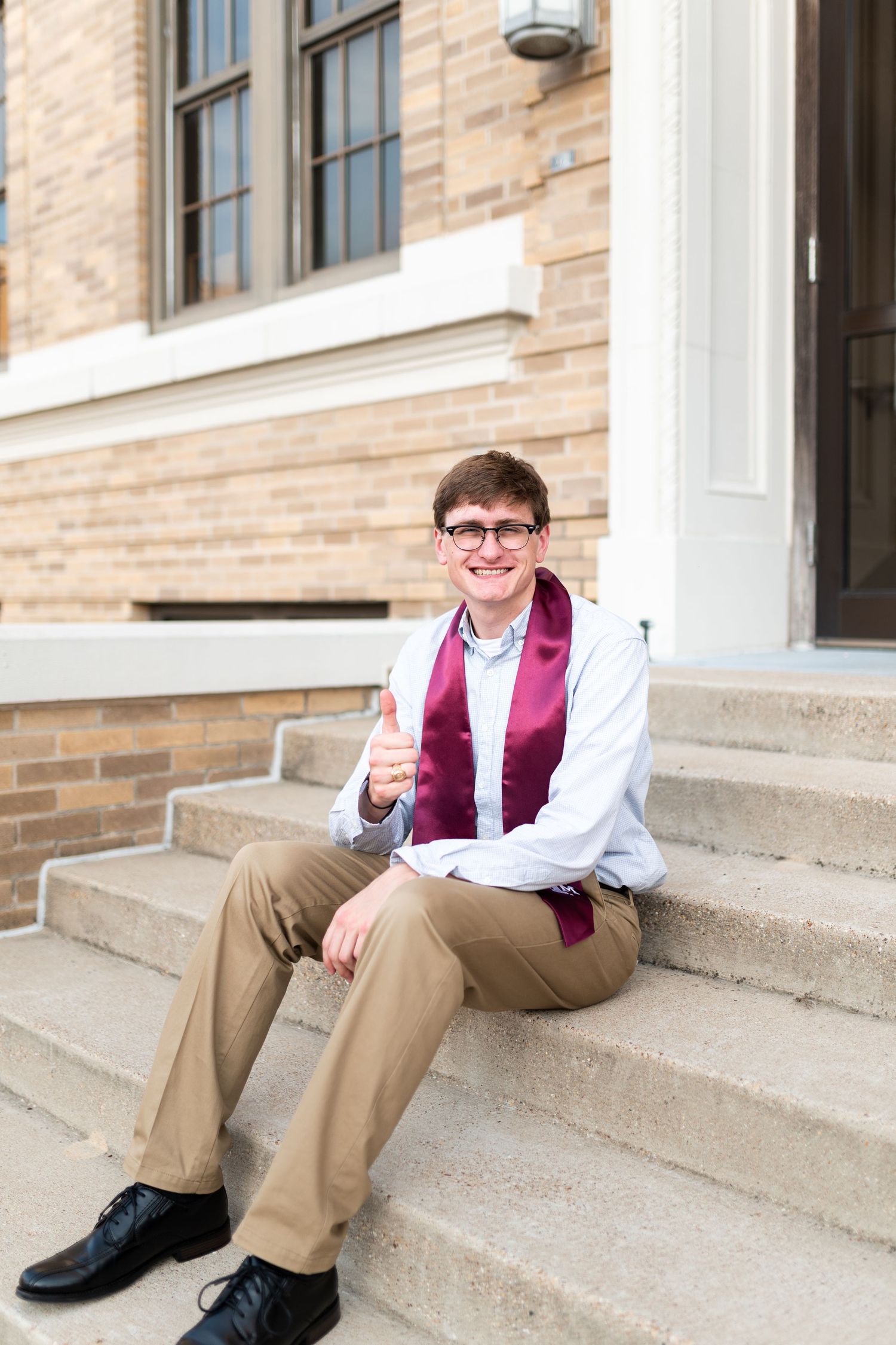 Young man in glasses giving thumbs up on steps wearing maroon Texas A&M graduation stole.