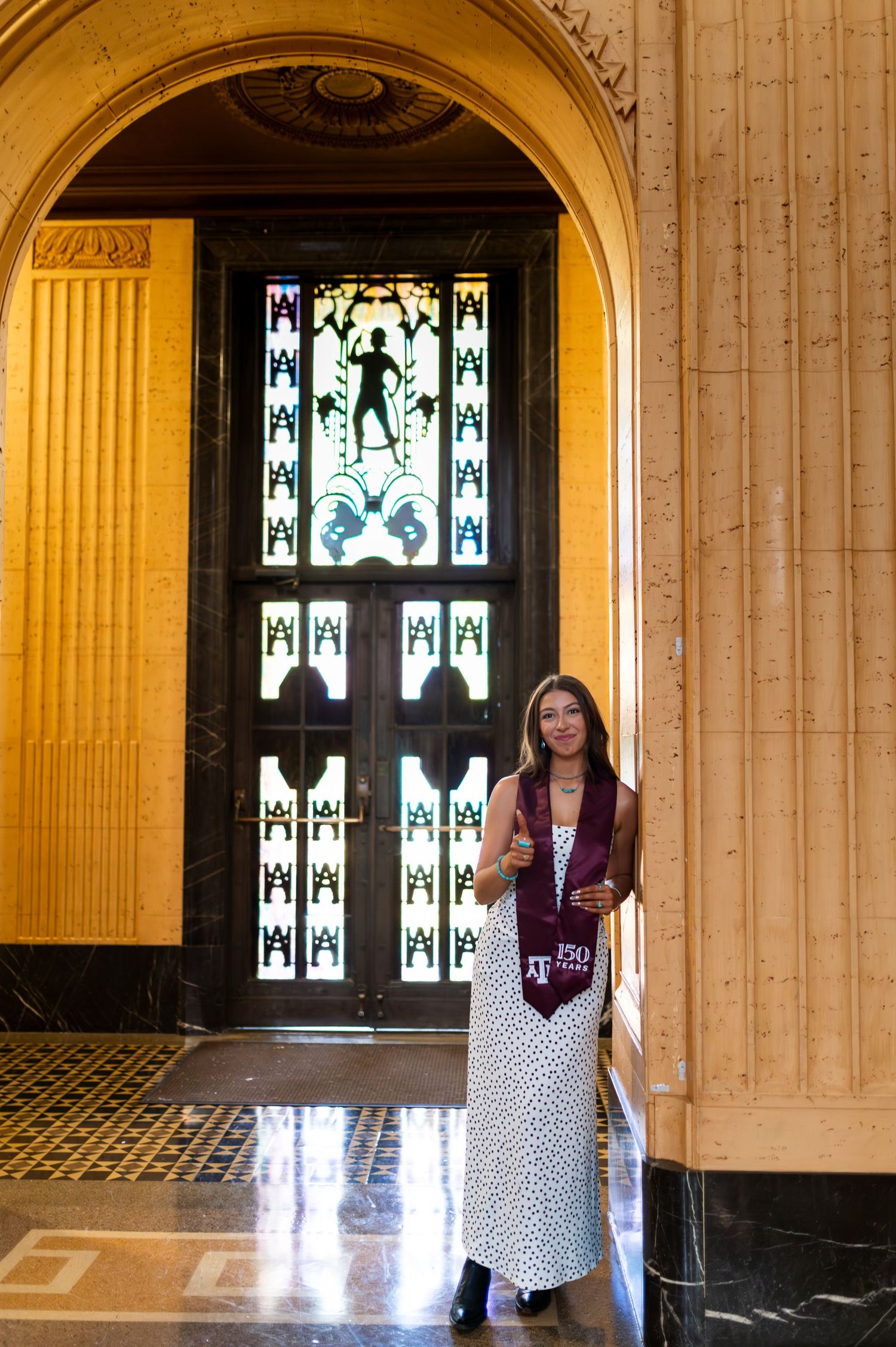 Texas A&M graduate stands before stunning stained glass window in elegant golden-hued building. Grad Photos at the Texas A&M Admin building