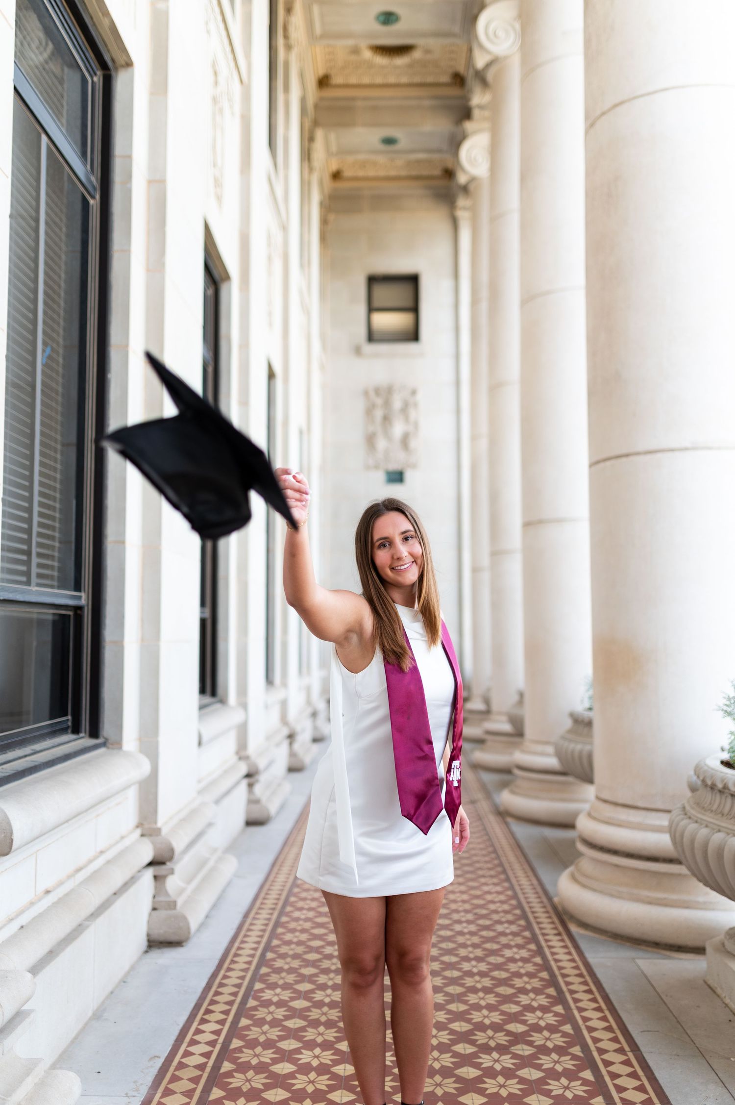 Female graduate in white dress and maroon stole tosses cap in columned hallway. Grad Photos at the Texas A&M Admin building