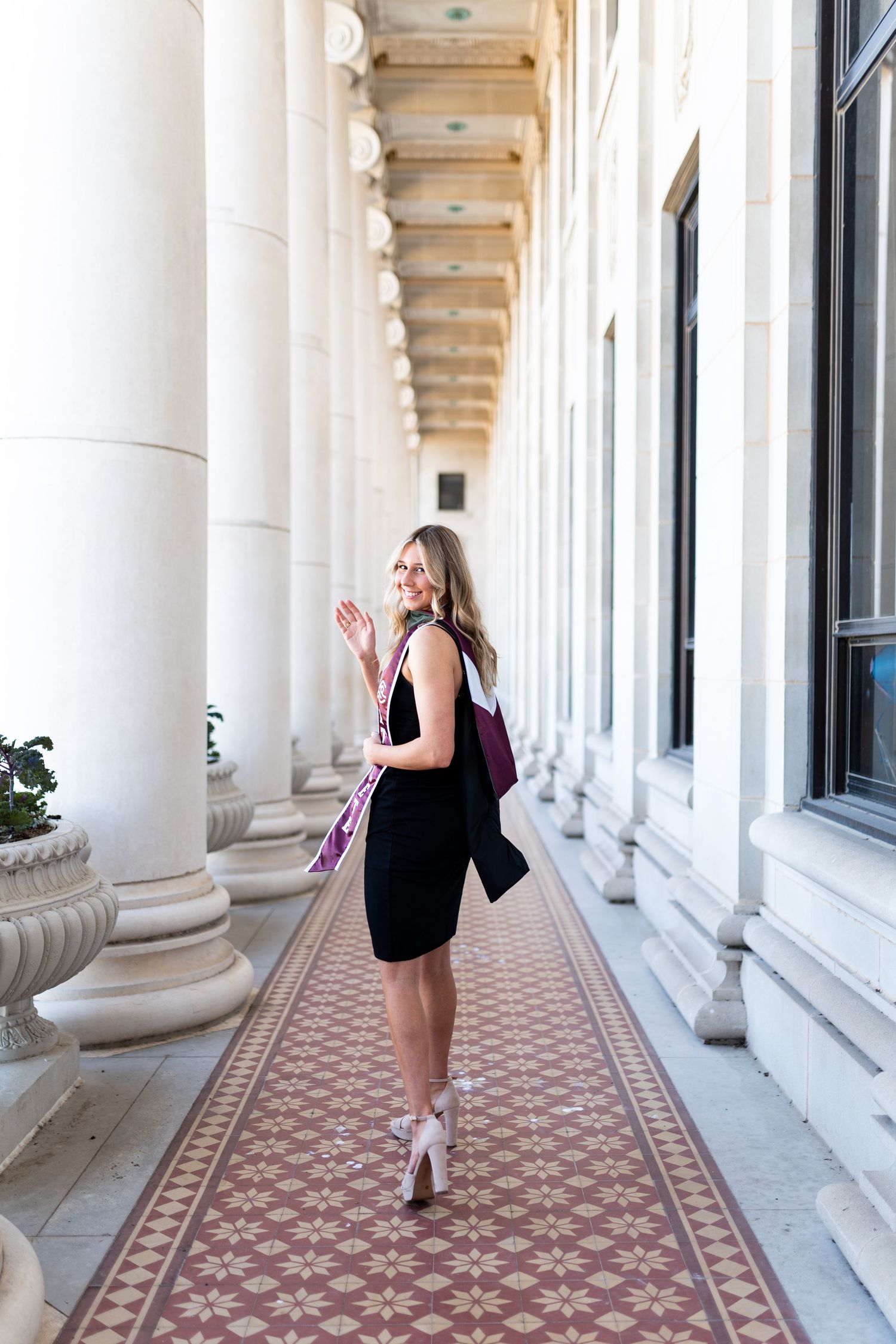 Blonde woman in black dress carrying graduation regalia walking through elegant columned corridor. Grad Photos at the Admin building