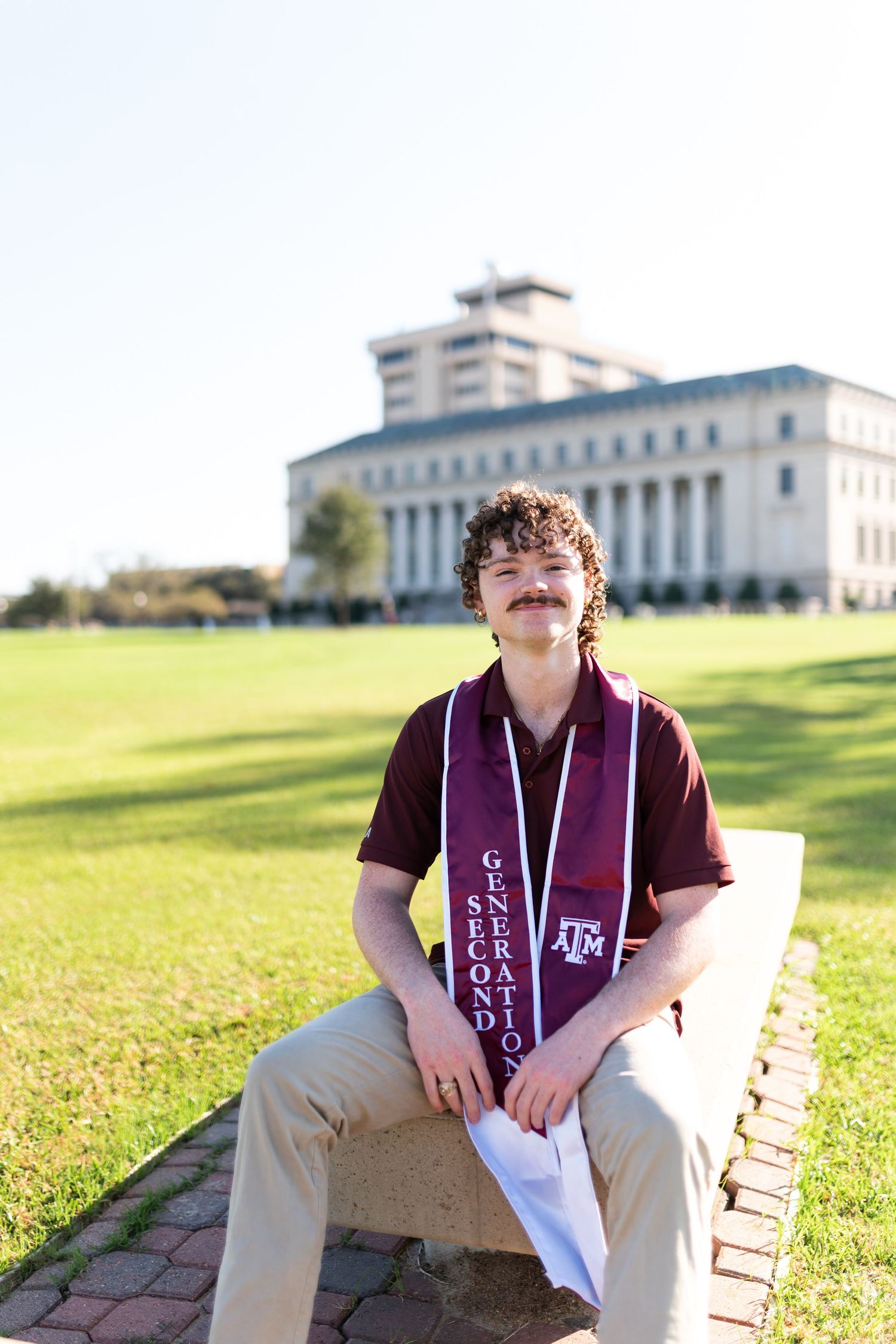 Young man with curly hair wearing Texas A&M second generation graduation stole seated on campus bench. Grad Photos at the Admin building