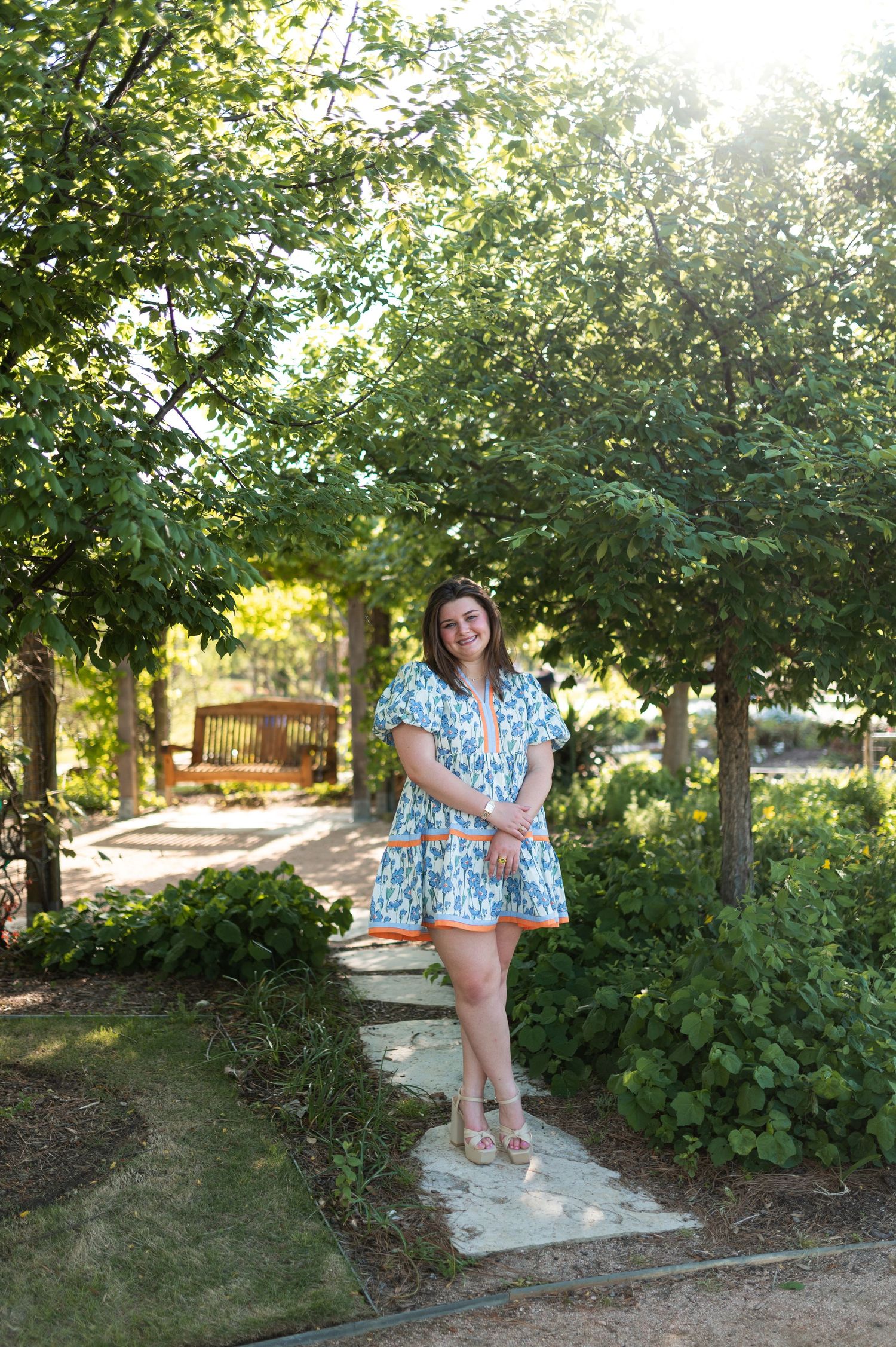 Young woman in blue floral dress standing on stone path in sunny green garden.