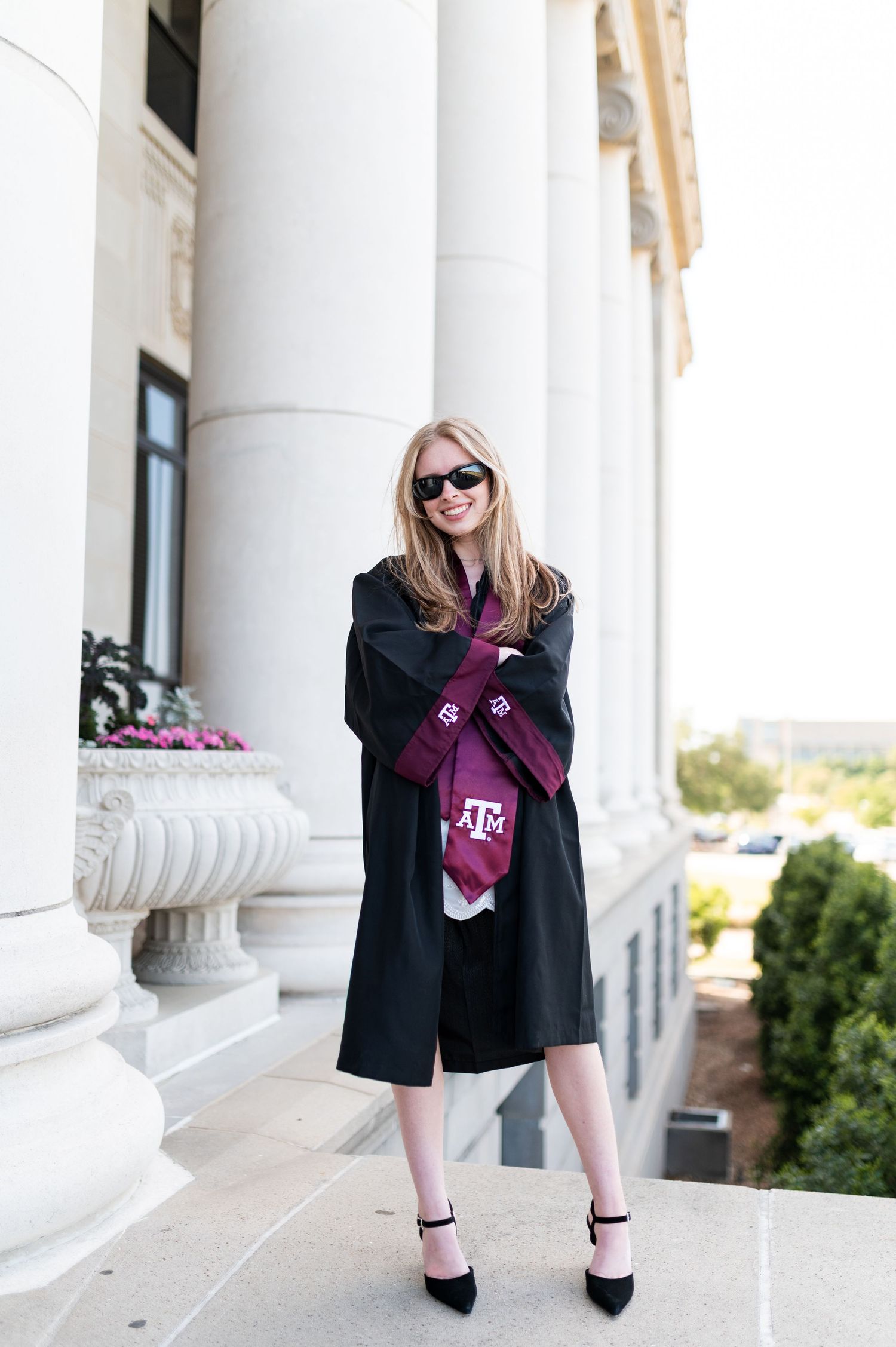 Texas A&M graduate in black gown and maroon stole poses confidently between white columns wearing sunglasses. Grad Photos at the Admin
