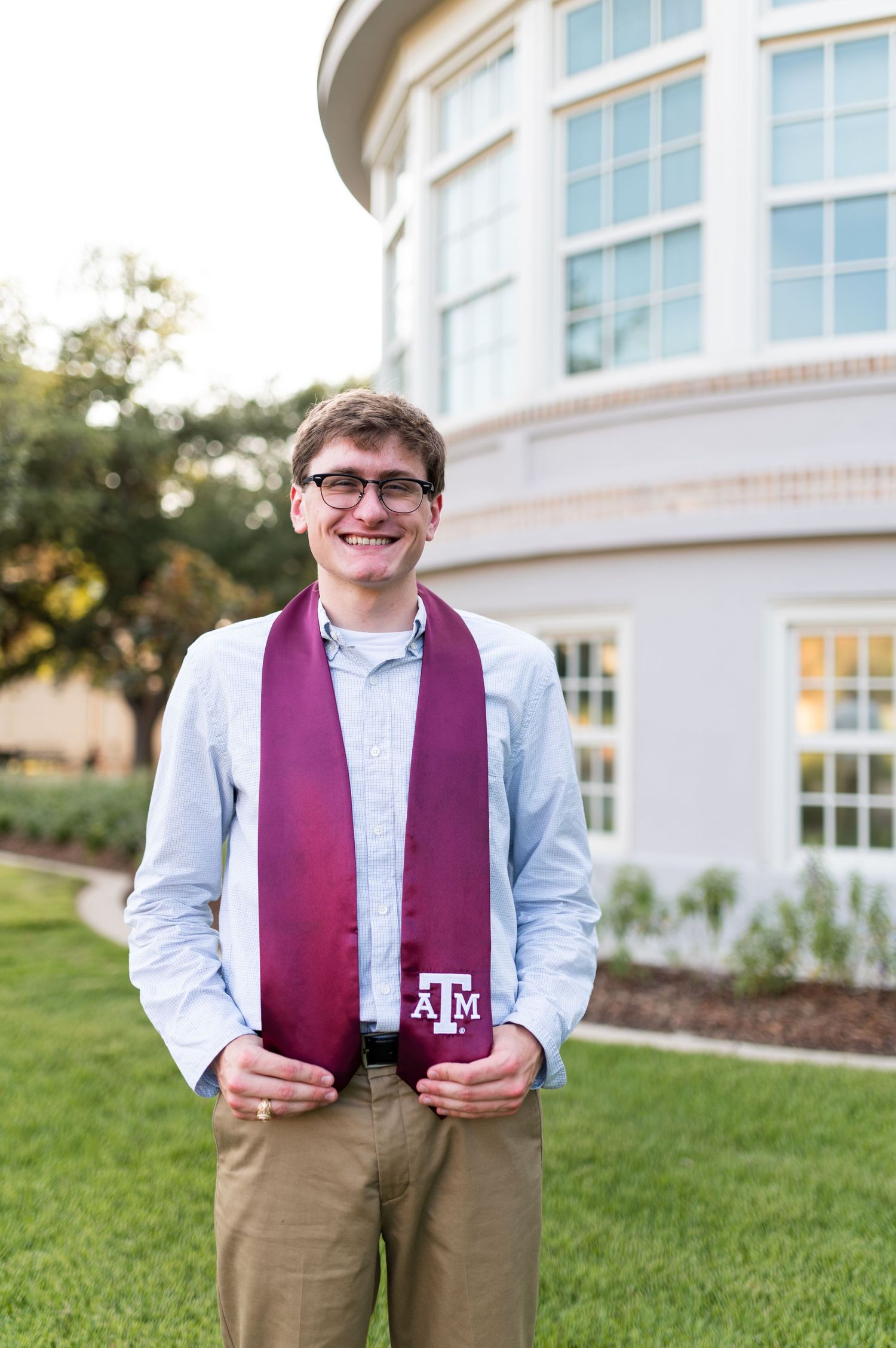 Smiling Texas A&M graduate poses outdoors wearing maroon Aggie stole over blue shirt with khaki pants.