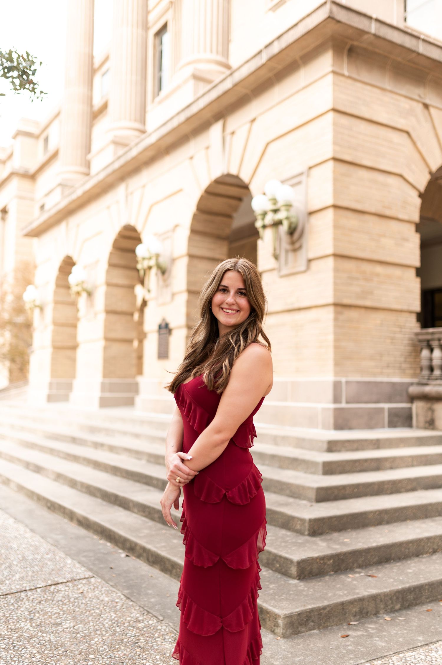 Smiling woman in tiered red formal gown poses on steps of a grand stone building at golden hour.