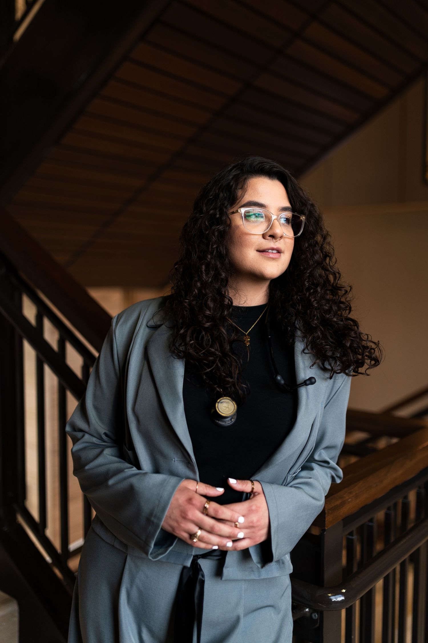 Young woman with curly hair and glasses in gray blazer poses thoughtfully on staircase indoors.