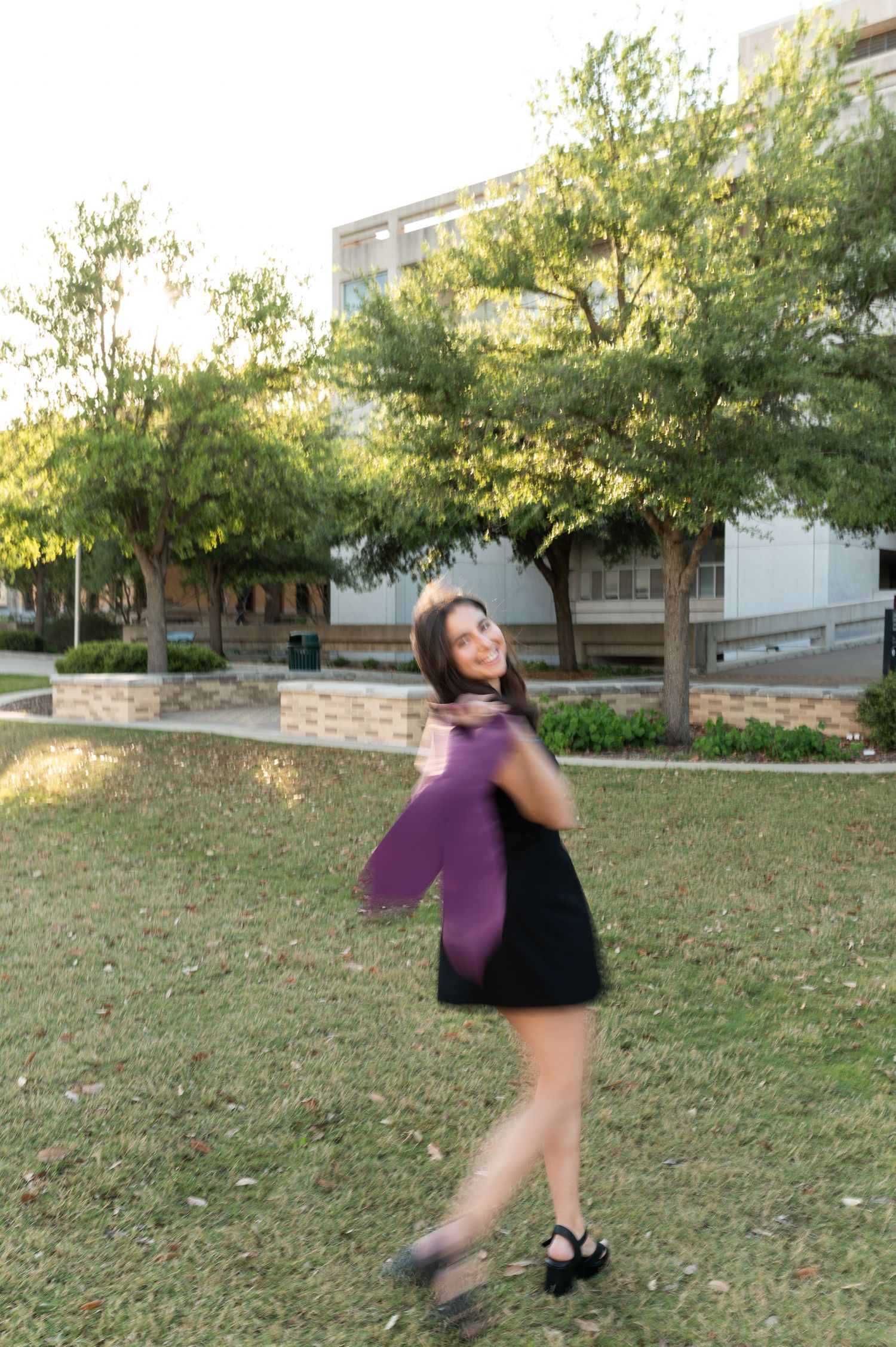 Smiling graduate in black dress twirling maroon stole in outdoor campus park setting.