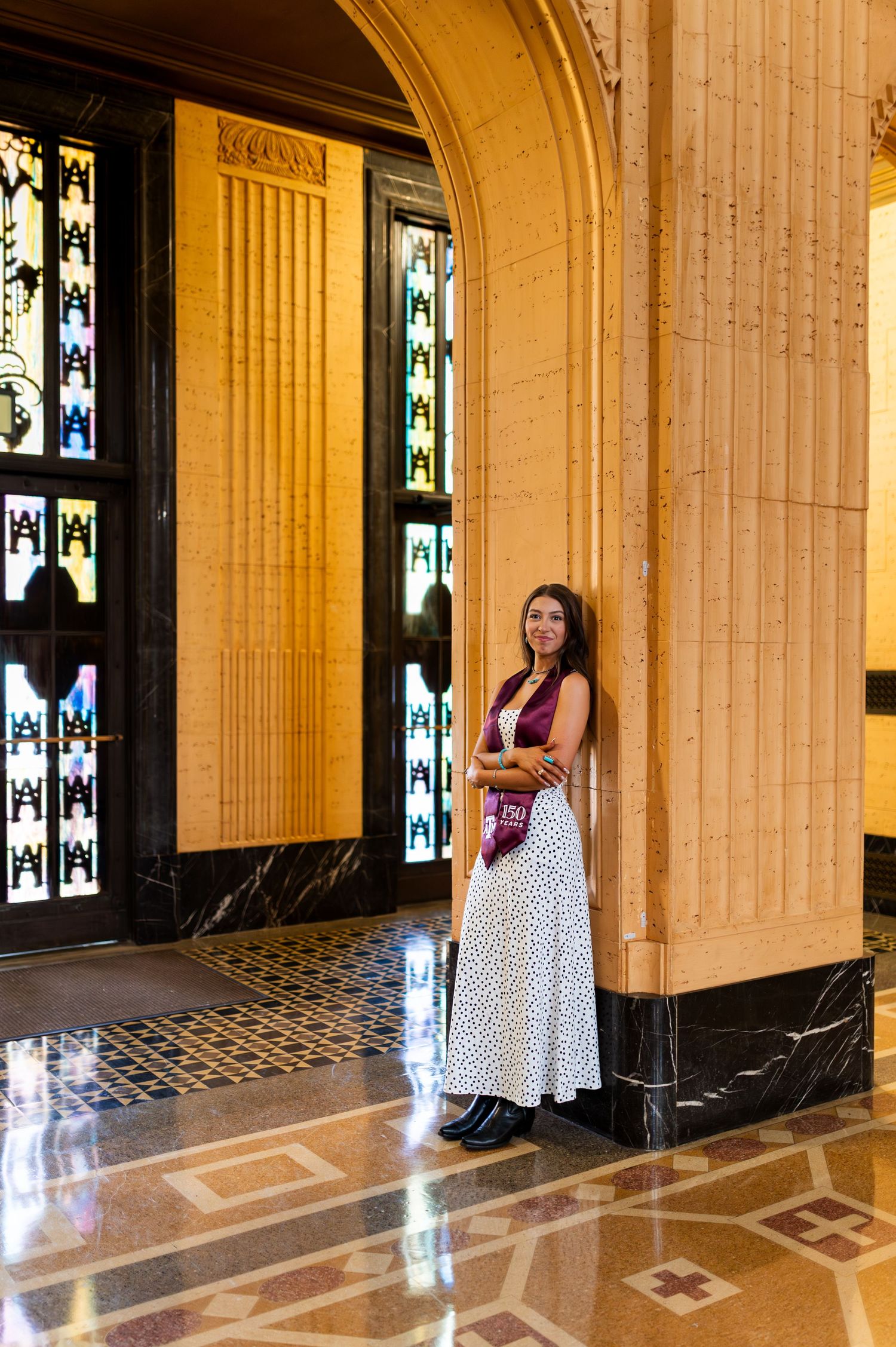 Graduate in polka dot maxi dress poses beside marble column in grand ornate building interior. Grad Photos at the Texas A&M Admin building