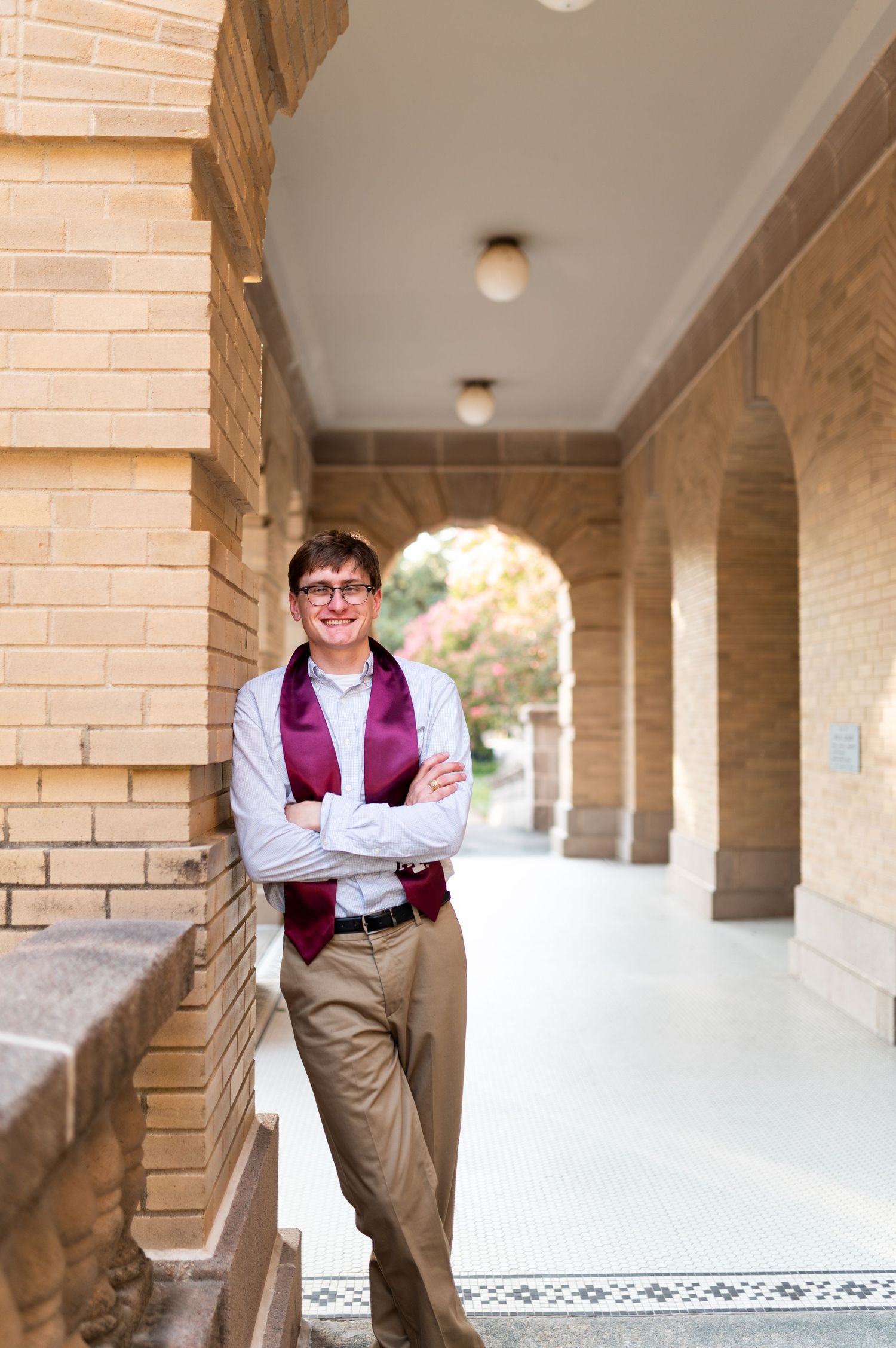 Male Texas A&M graduate with maroon stole poses in brick archway corridor on campus.
