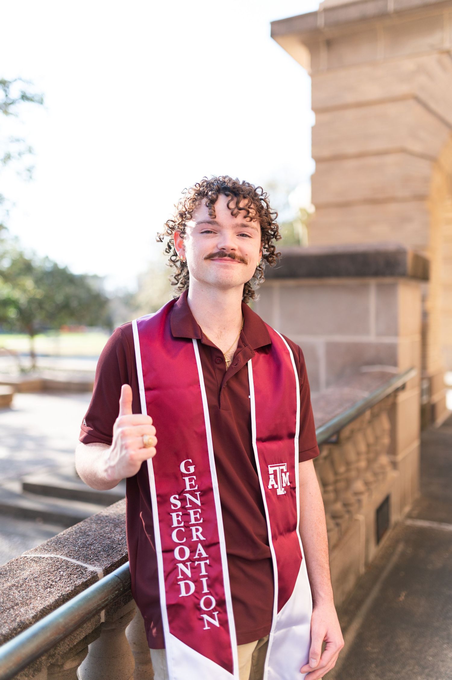 Curly-haired graduate holds thumbs up wearing Second Generation Texas A&M stole.