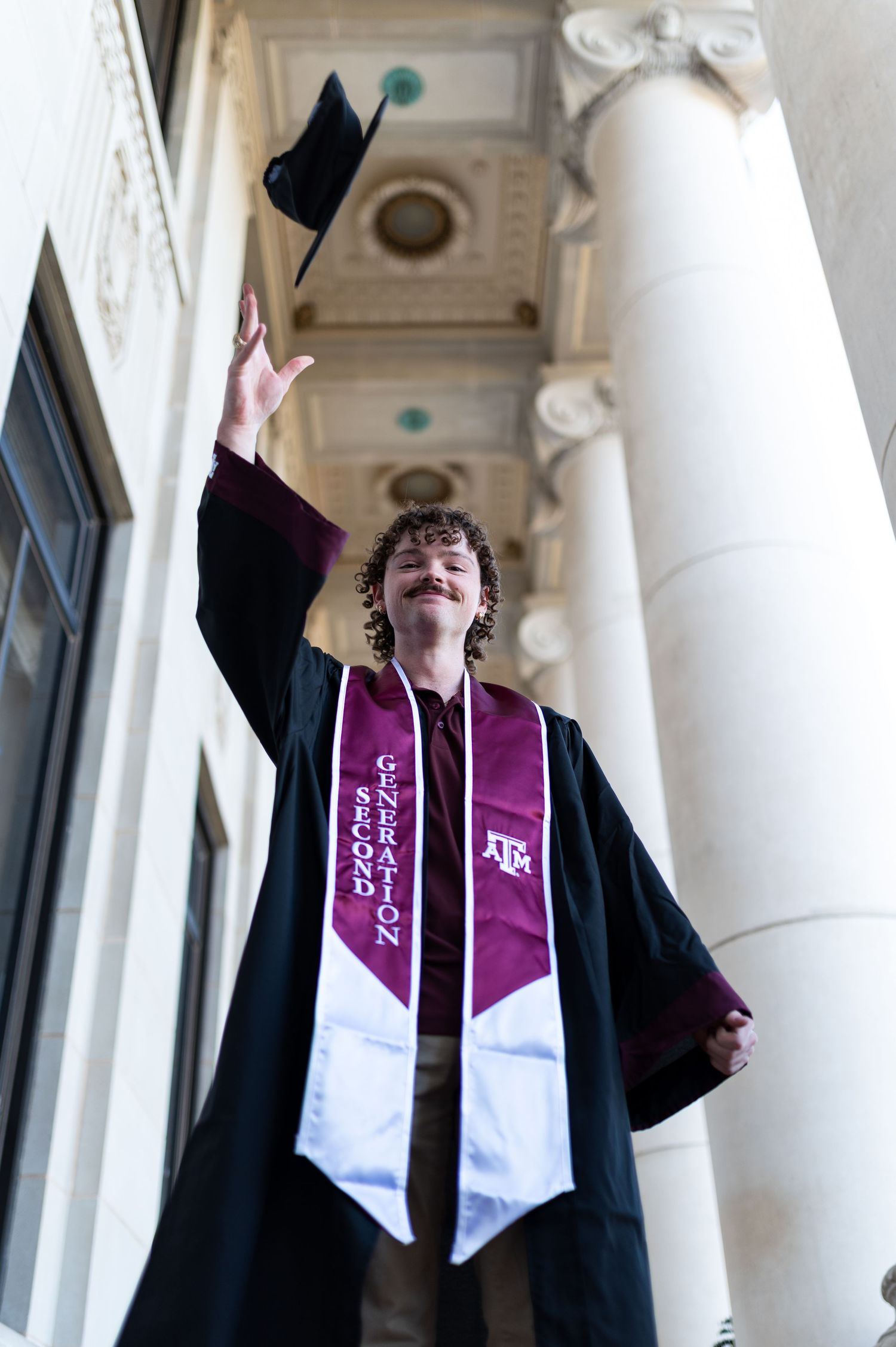 Graduate in black cap and gown tossing cap in air wearing Texas A&M second generation stole. Texas A&M Grad Photos at the Admin building