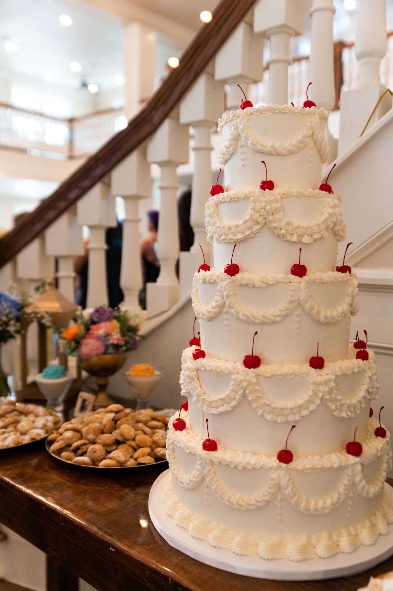 Five-tier white wedding cake decorated with red cherries and ruffled frosting displayed near a grand staircase.