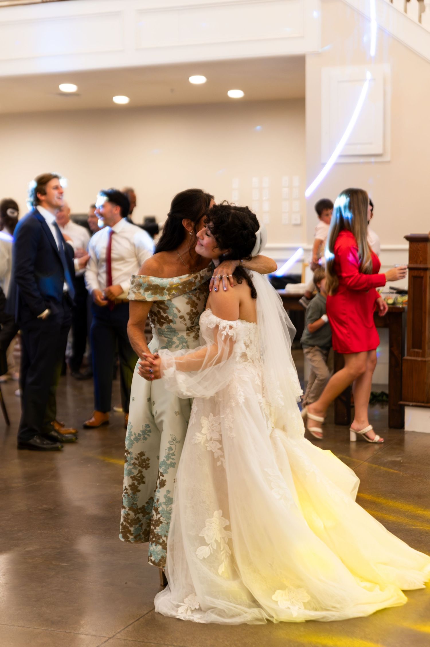 Bride in white lace gown shares heartfelt embrace with guest in floral dress on dance floor.
