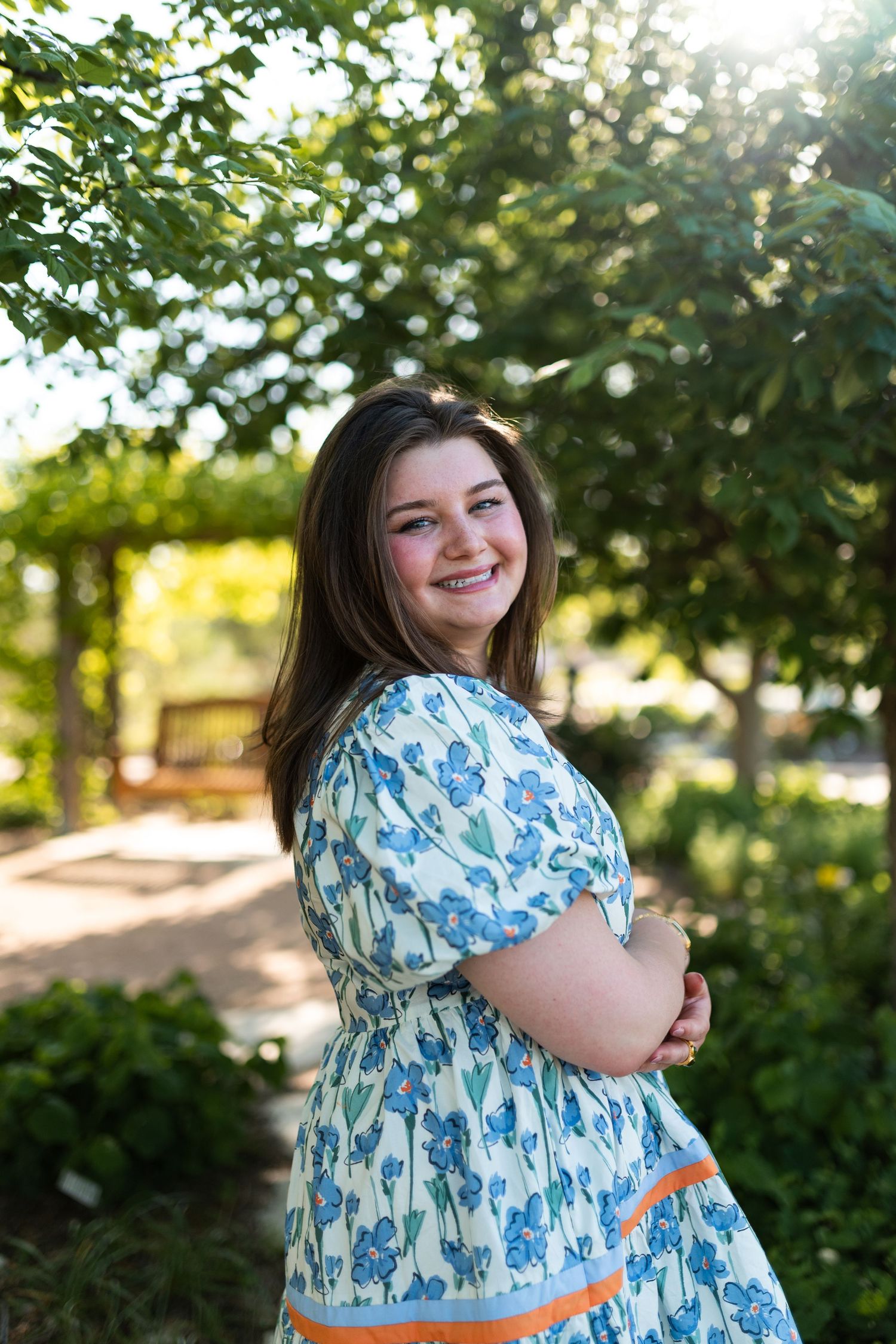 Smiling young woman in blue floral dress standing in lush green garden setting.