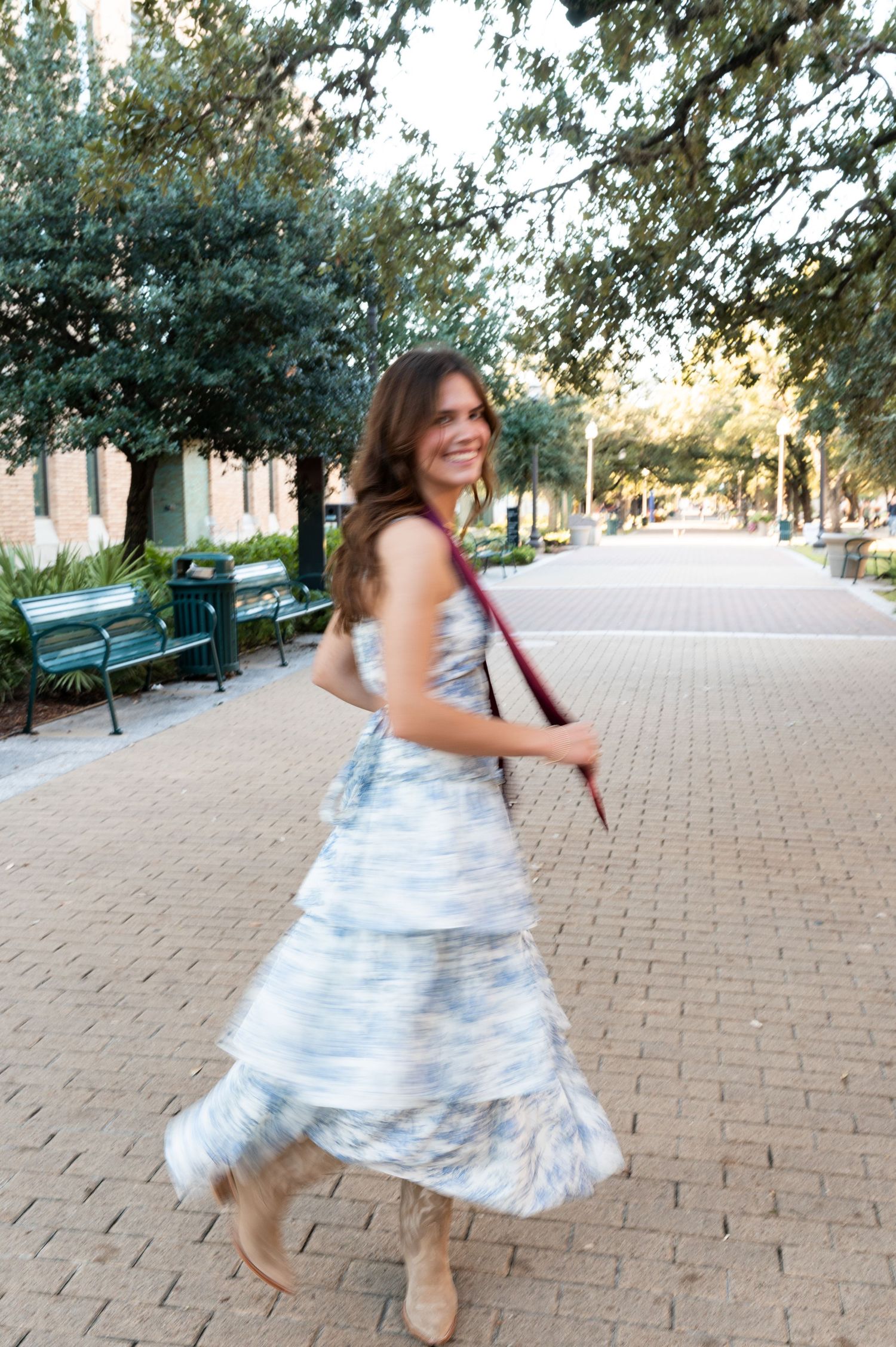 Joyful Texas A&M graduate spins in blue floral dress with maroon stole on tree-lined brick pathway.