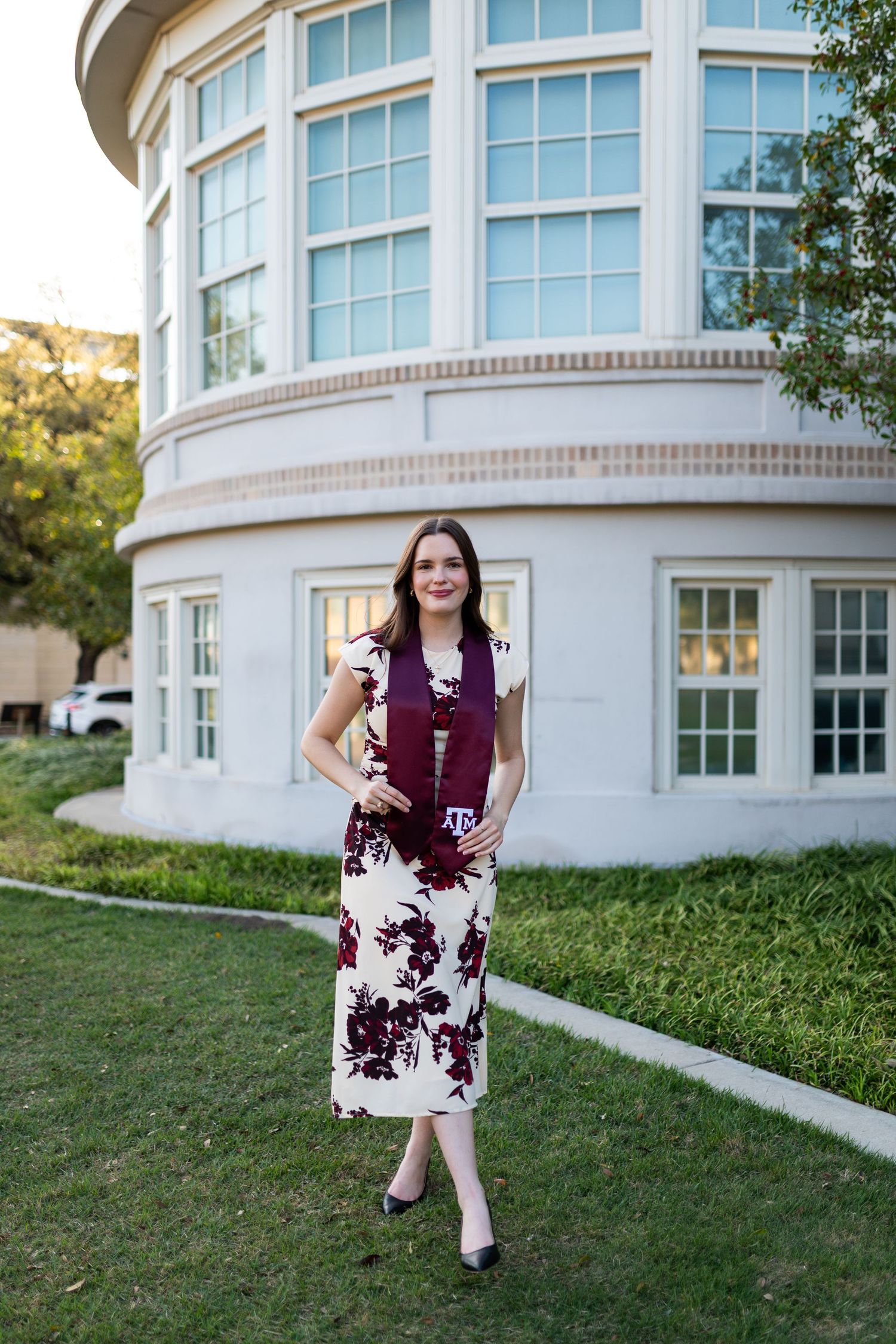 Female Texas A&M graduate poses in floral dress with maroon stole on green lawn near campus building.