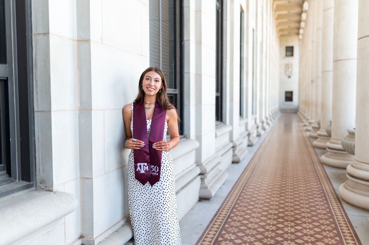Woman in polka dot dress with Texas A&M maroon stole stands in sunlit colonnade. Grad Photos at the Admin building