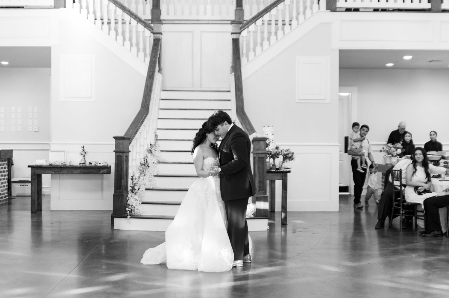 A bride and groom share their first dance in black and white photo beneath an elegant staircase at their reception.