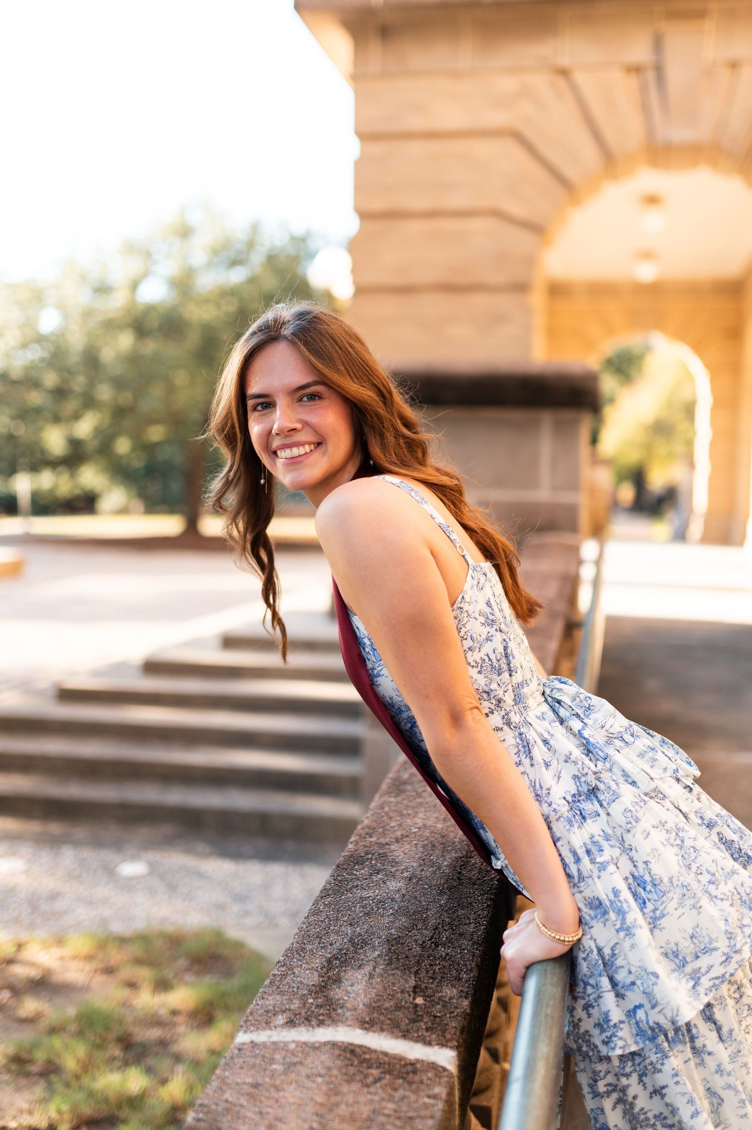 Smiling Texas A&M graduate in blue floral dress leans on stone railing near historic campus archway.